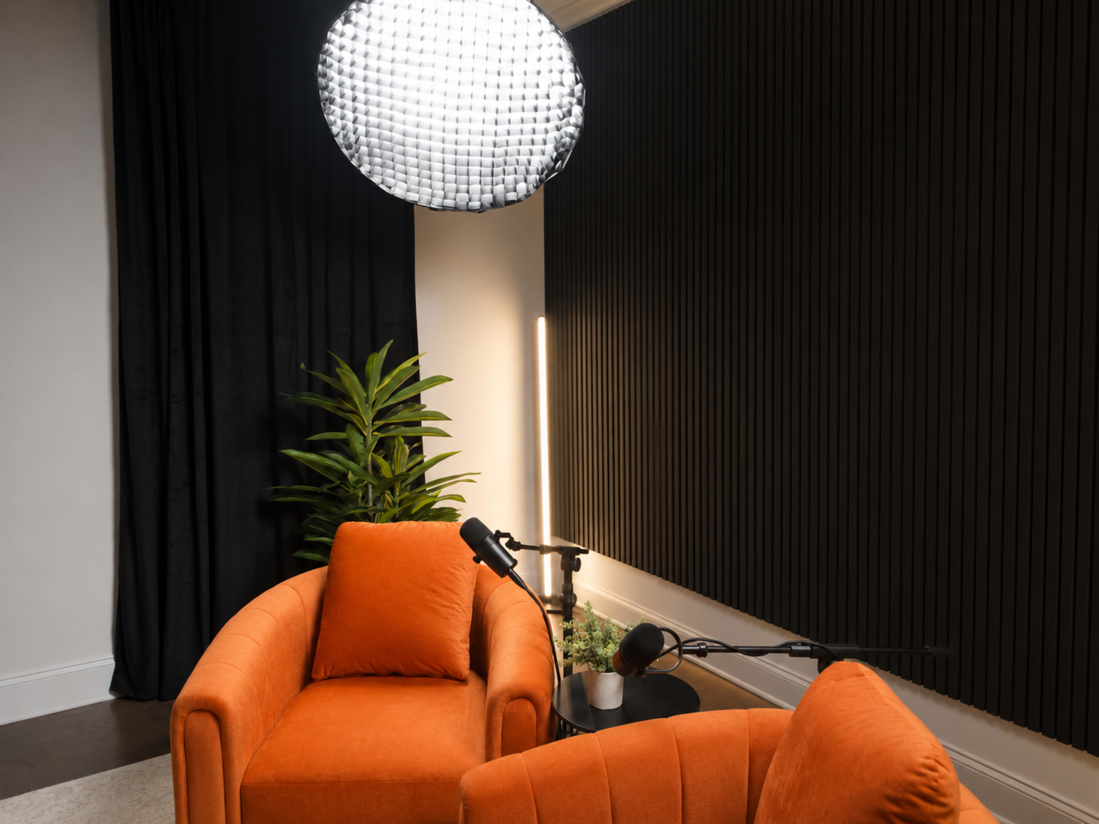 The image shows a modern podcast studio featuring two orange armchairs, a large pendant light, and a microphone setup against a dark, textured wall.