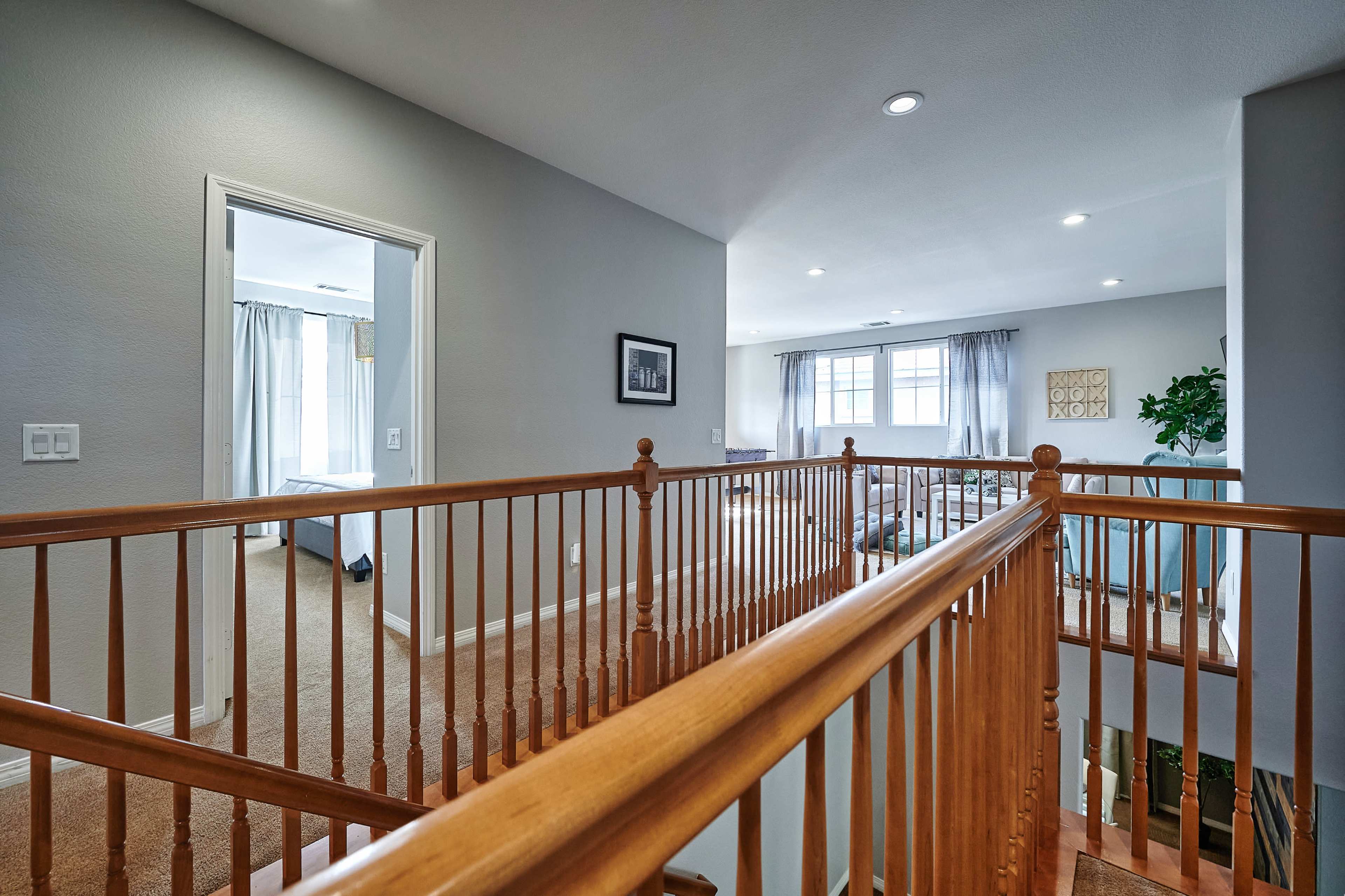 A well-lit second-floor hallway with wooden railings, leading to two rooms with large windows.