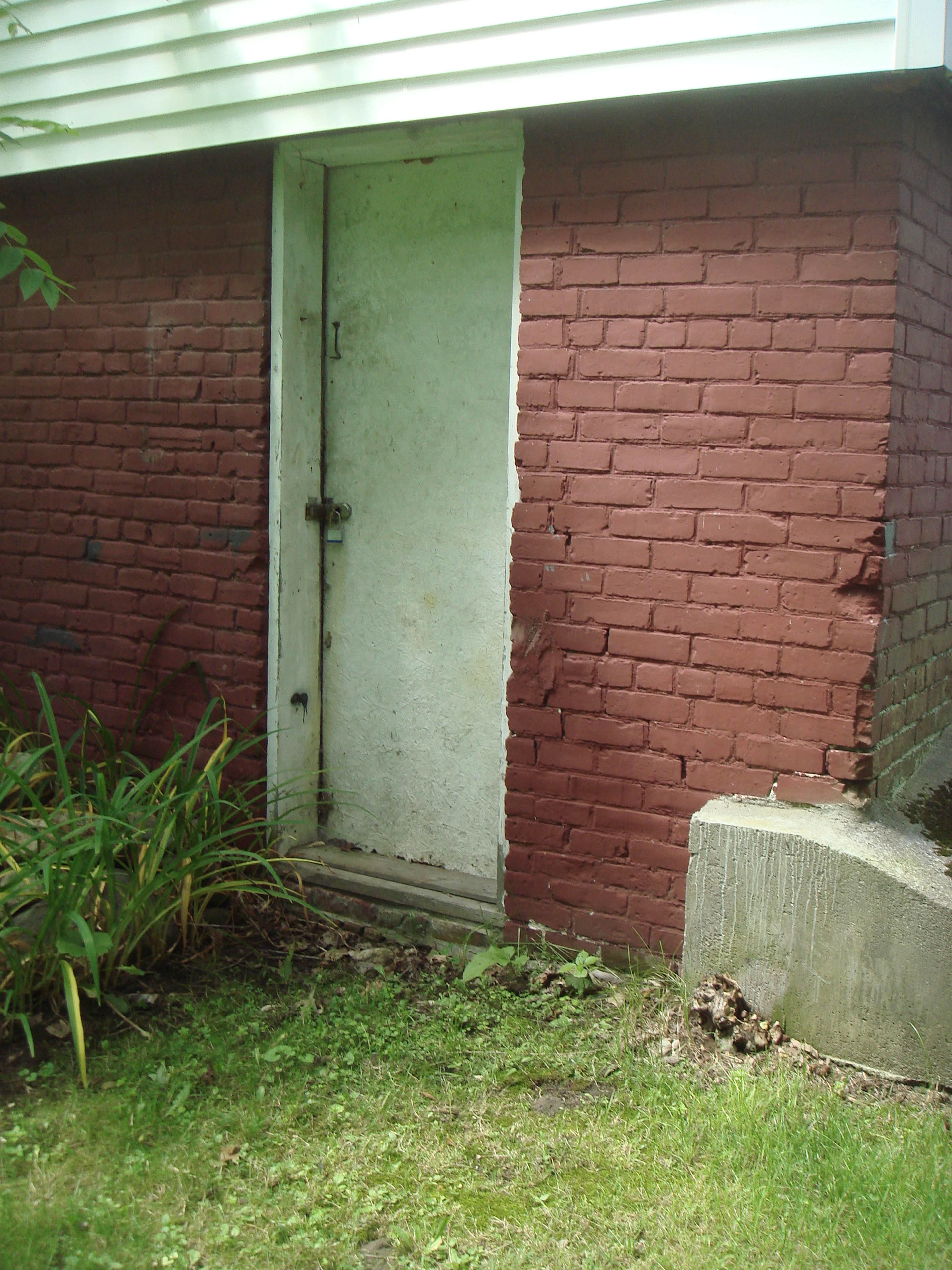 A weathered, white door set into a brick wall, with a patch of brown bricks on one side and greenery in front.