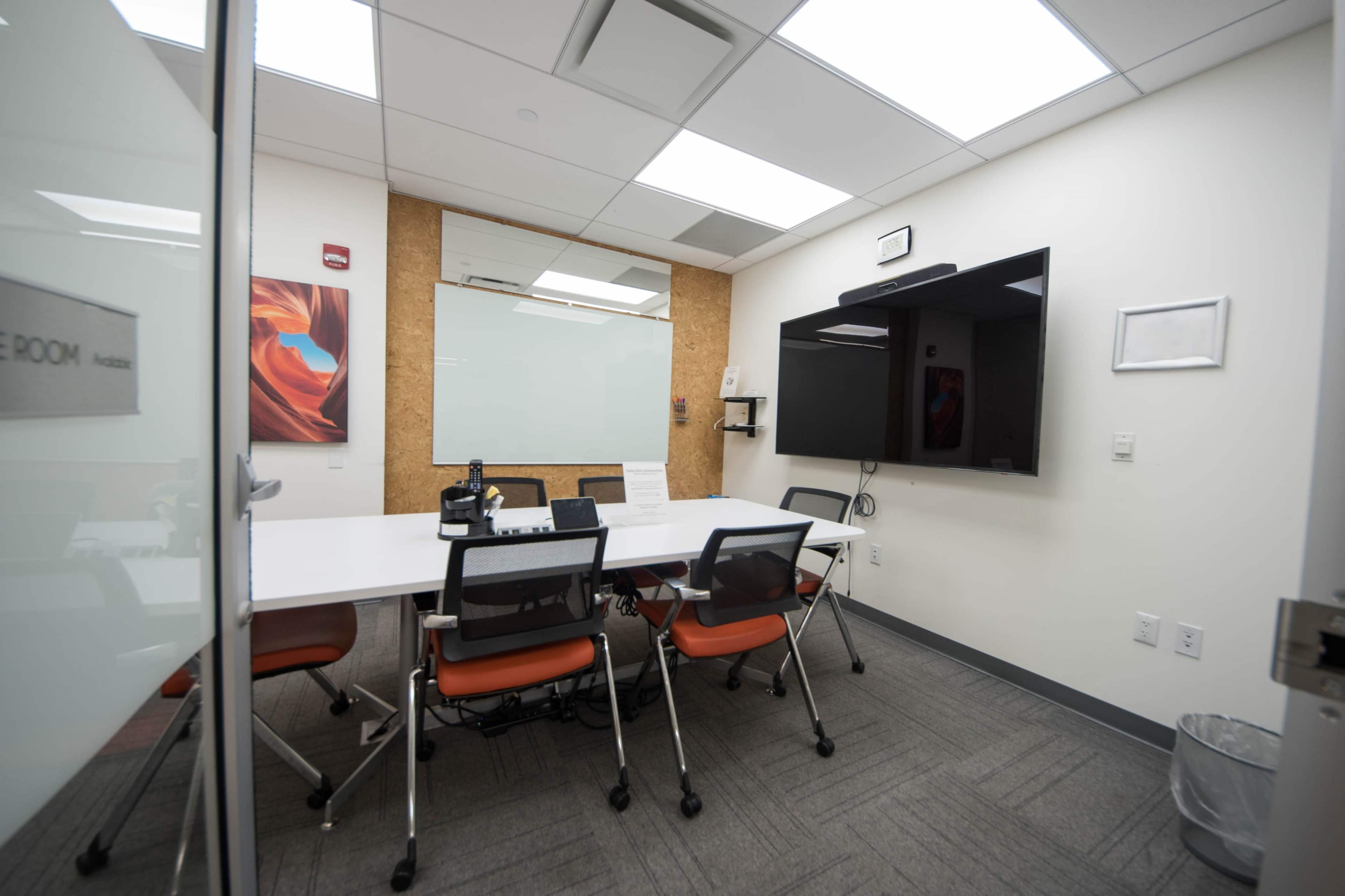 A small conference room is equipped with a whiteboard, a large screen, and a meeting table surrounded by orange chairs.