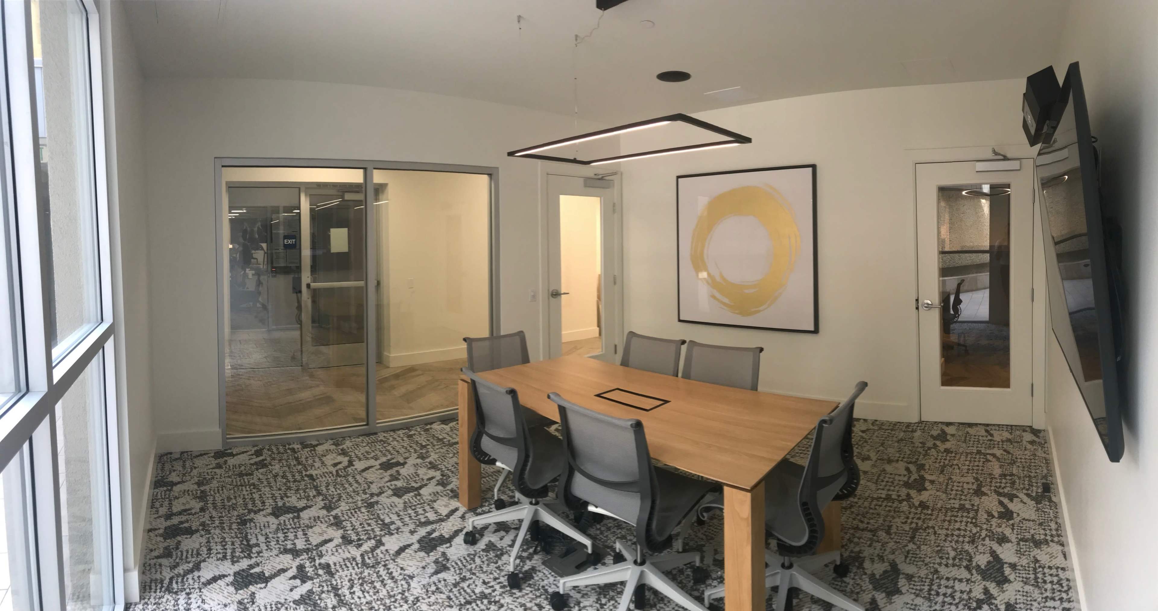 A modern conference room features a wooden table surrounded by gray chairs, with large windows and a yellow circular artwork on the wall.