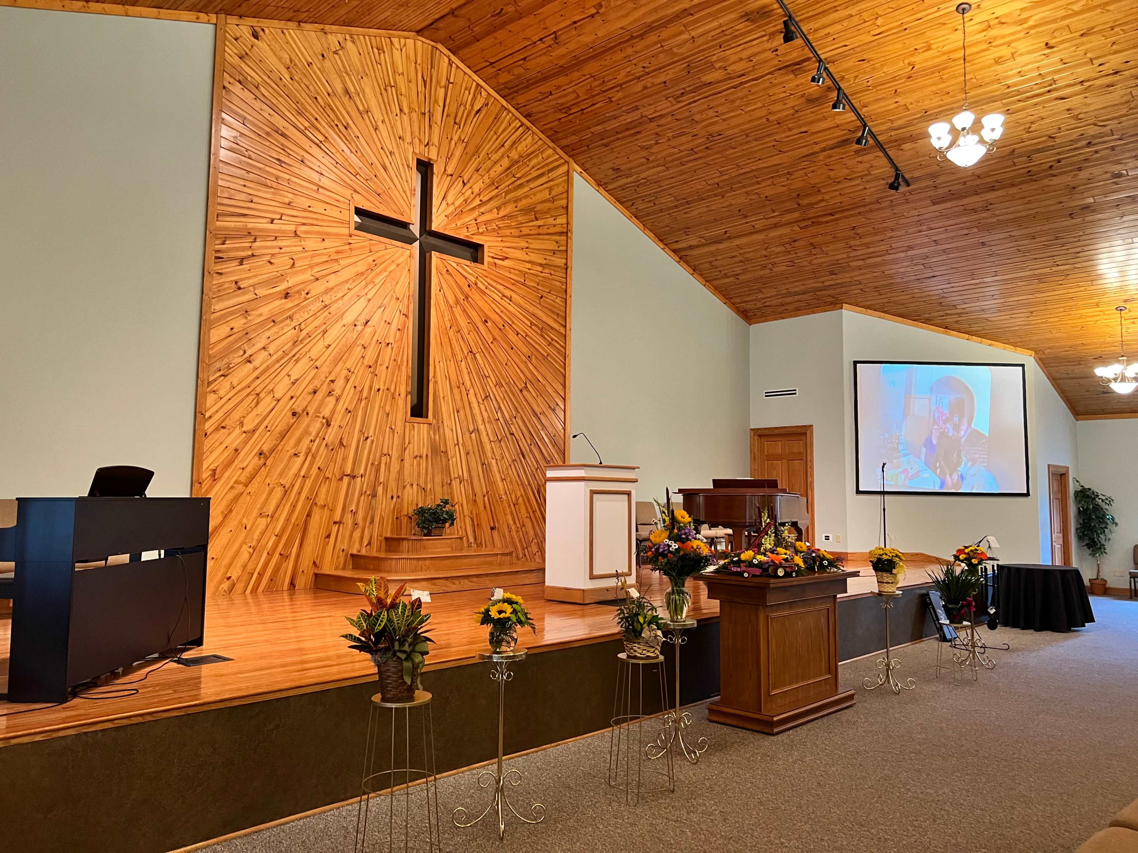 An interior view of a church with a wooden cross on the wall and a stage area featuring floral arrangements and a large screen displaying a person.