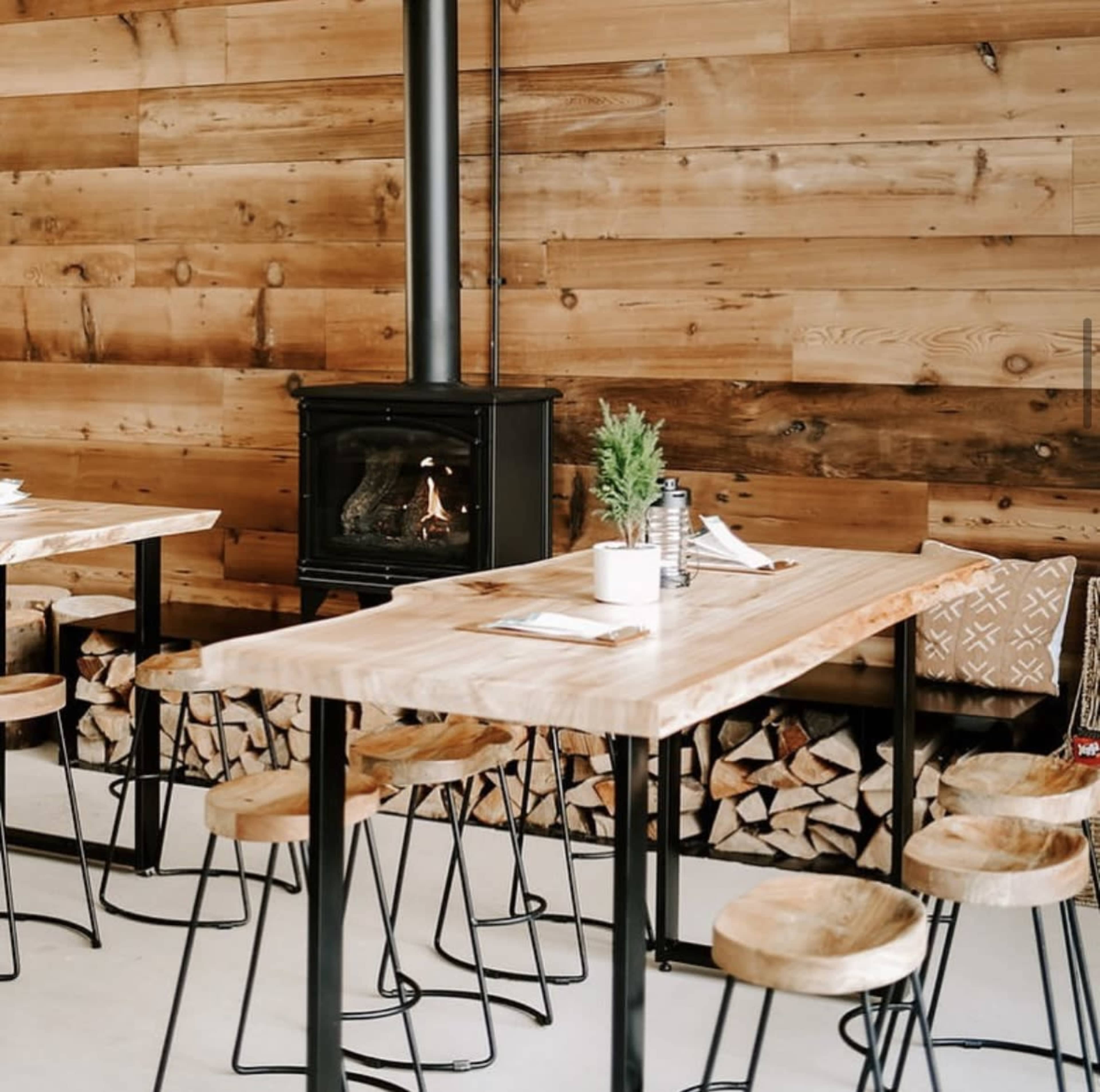 A rustic dining area with wooden tables and stools, a black wood stove, and a stacked firewood display against a wooden wall.