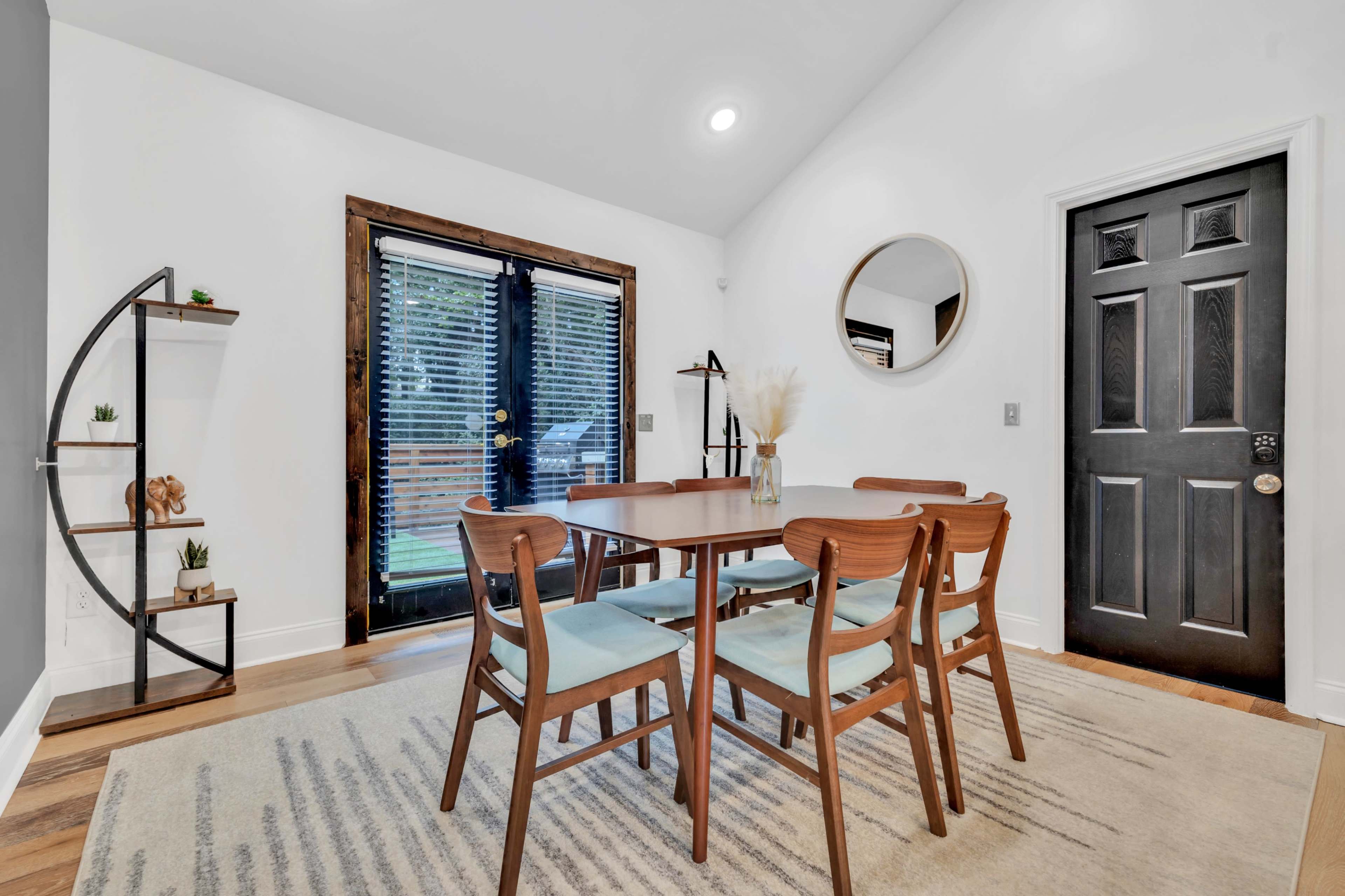 A modern dining area features a wooden table surrounded by four chairs, a circular mirror on the wall, and a black door leading outside.