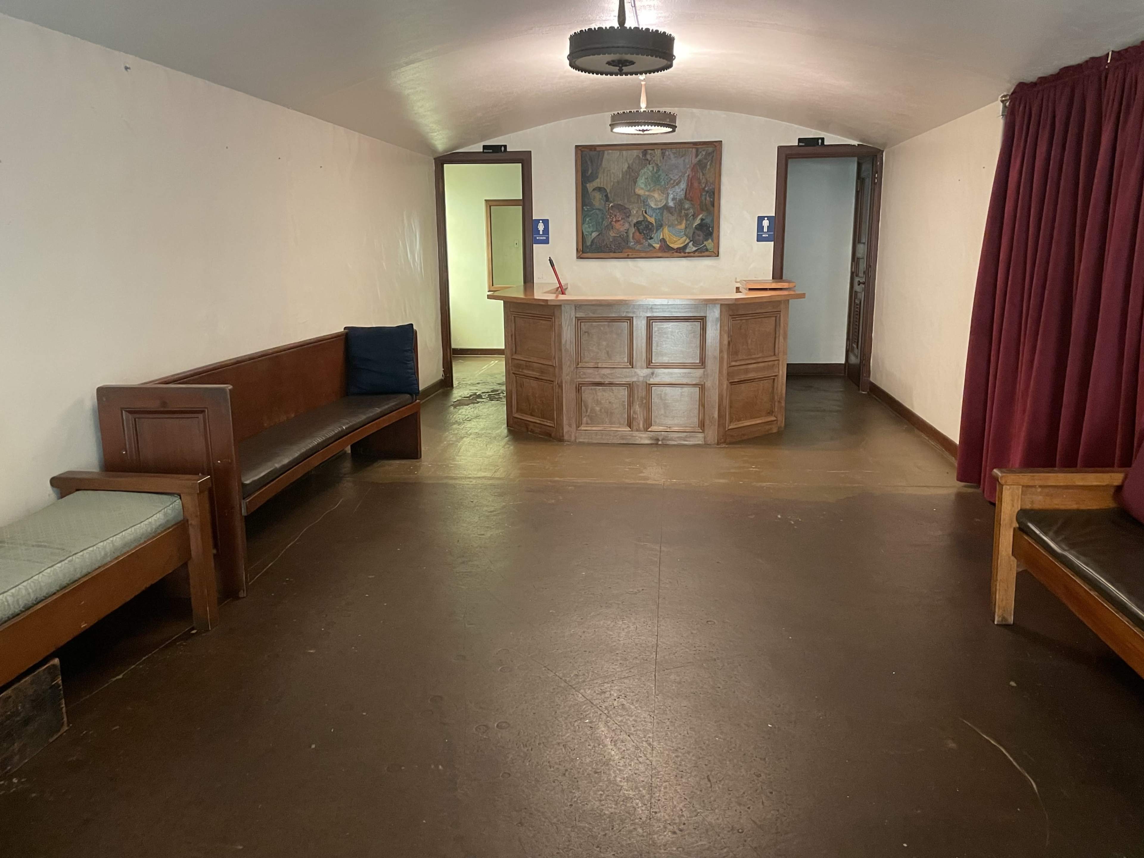 The image shows a small lobby area with wooden benches, a reception desk, and two doorways leading to other rooms, complemented by a wall painting and red curtains.