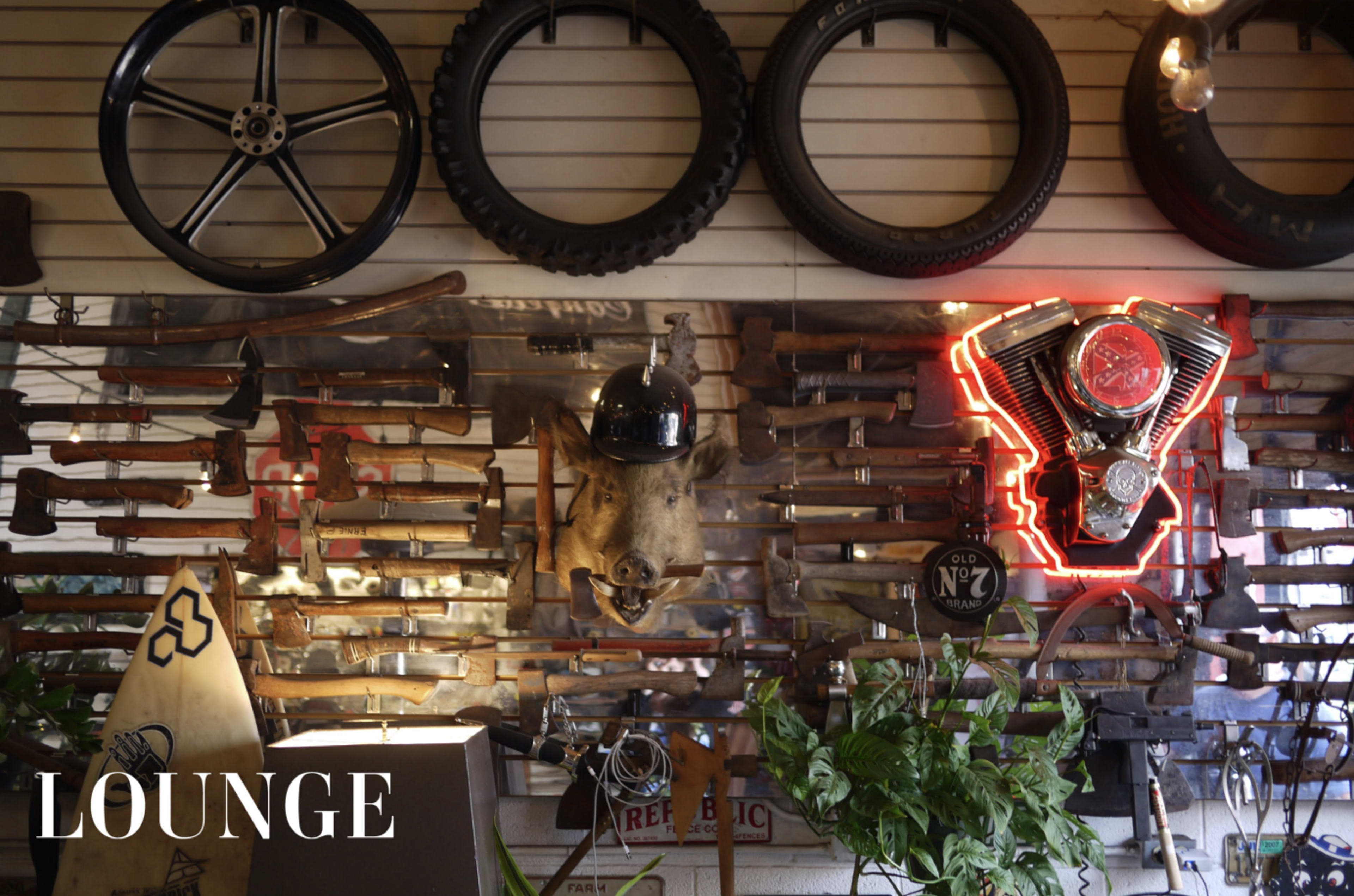 The image features a wall adorned with various antique tools, a motorcycle engine, a helmet, and decorative neon signage, all set against a backdrop of tires and plants.