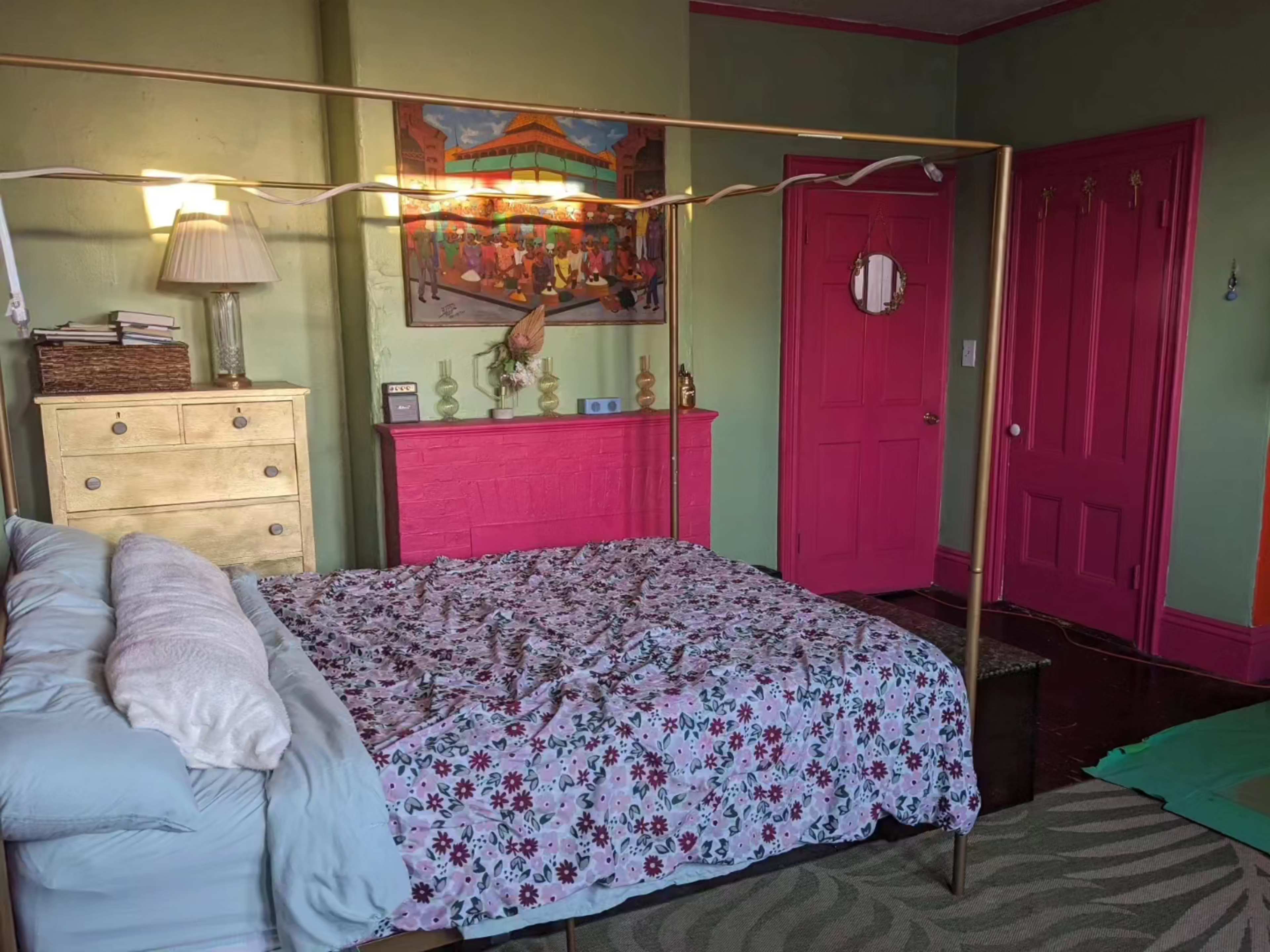 A bedroom featuring a gold four-poster bed with a floral bedspread, a wooden dresser, a brightly colored pink door, and green walls adorned with a large painting.