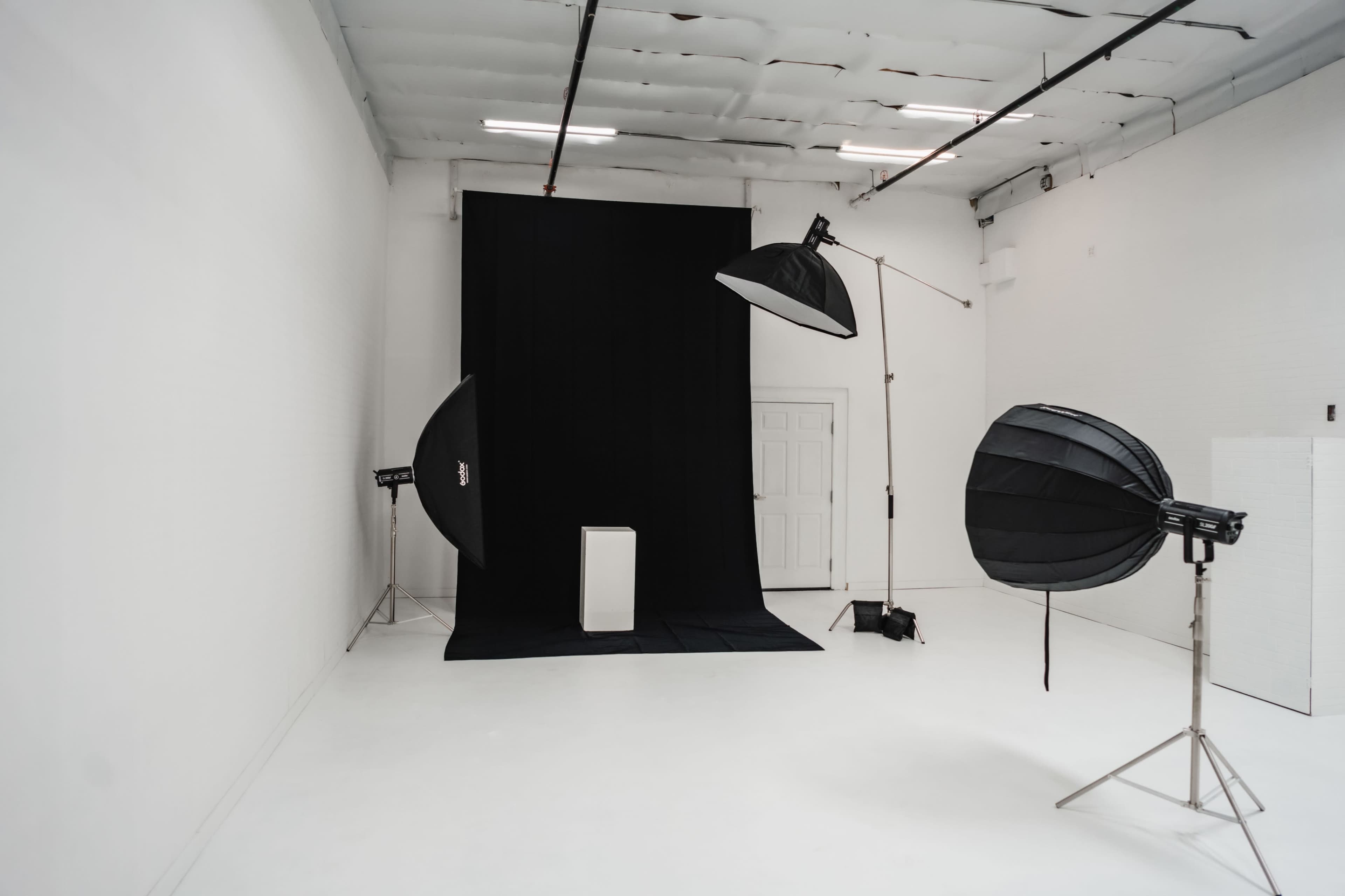 A photo studio features a black backdrop with a white pedestal and two large softbox lights positioned on either side.