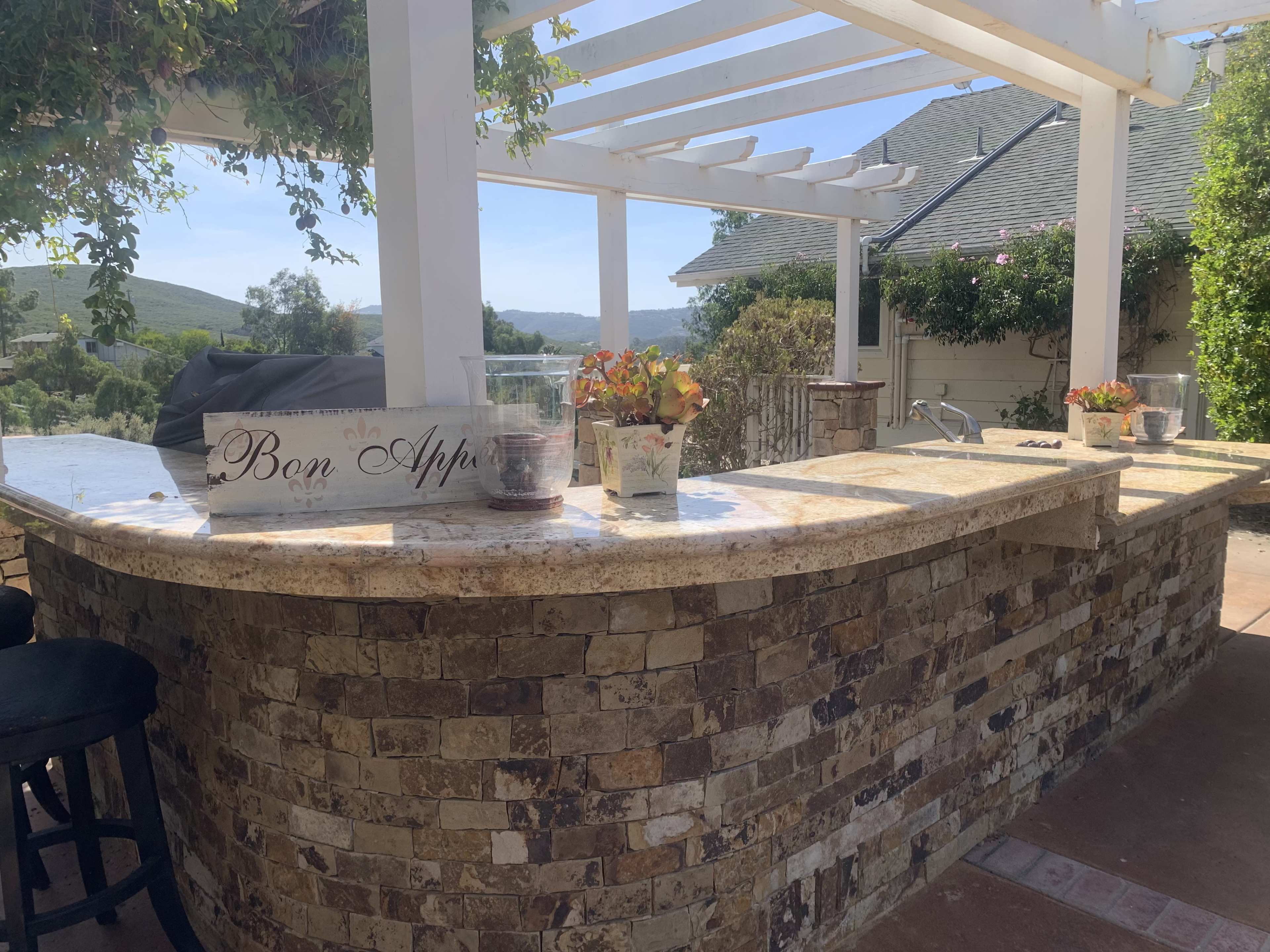 An outdoor kitchen area with a stone countertop and bar seating, surrounded by greenery and a clear sky.