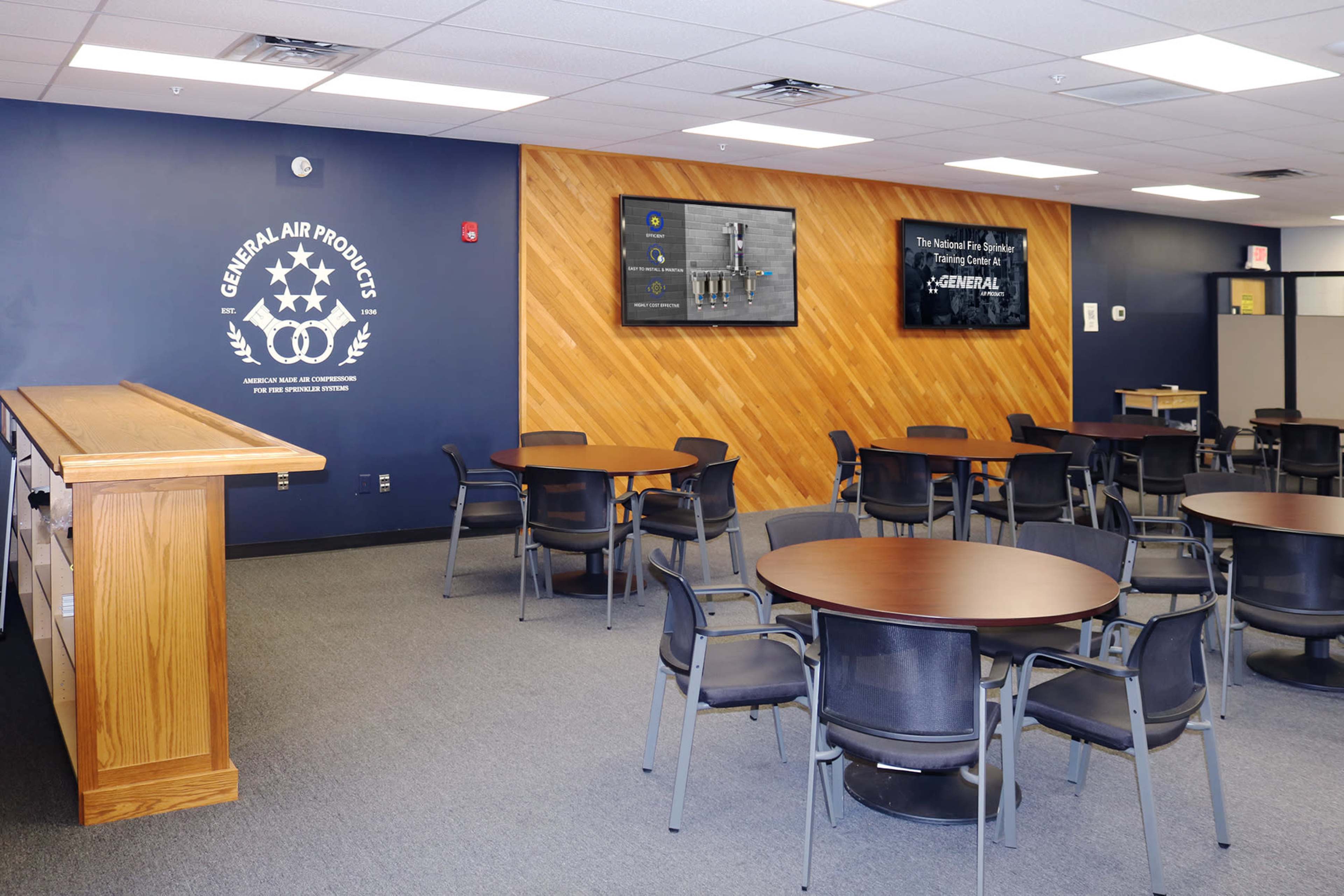 A modern office lounge area with wooden accents, circular tables, and chairs, featuring display screens on the wall.