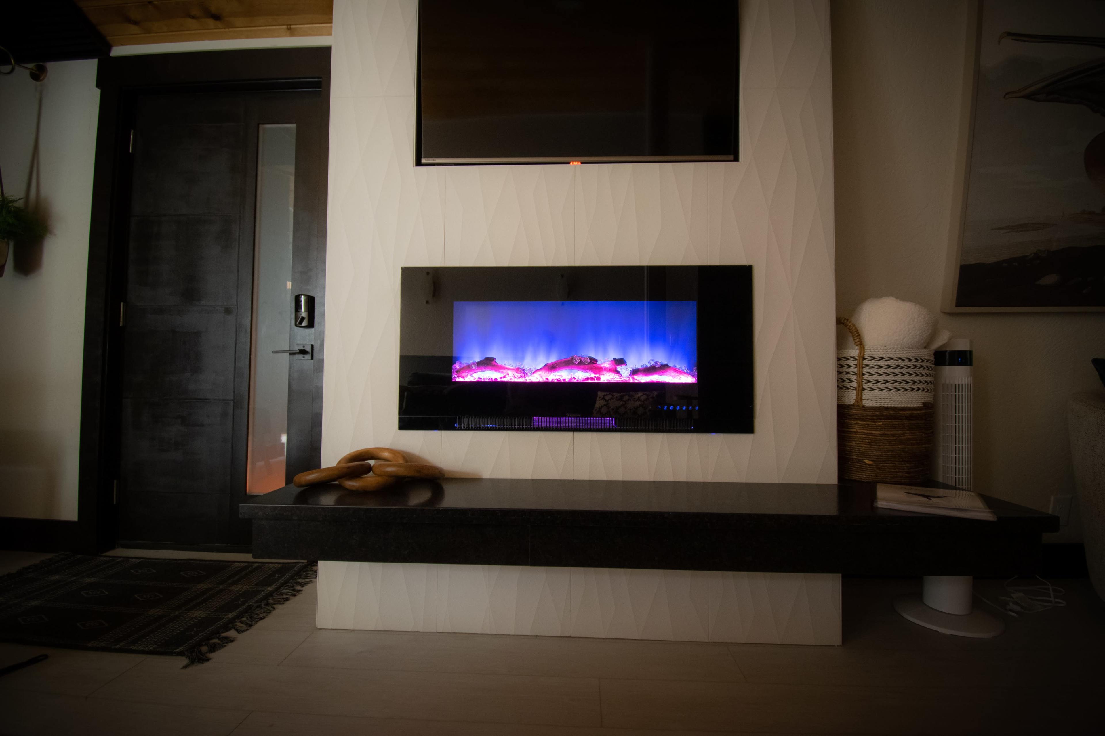 The image features a modern electric fireplace integrated into a textured white wall, with a dark shelf underneath and a television mounted above it.