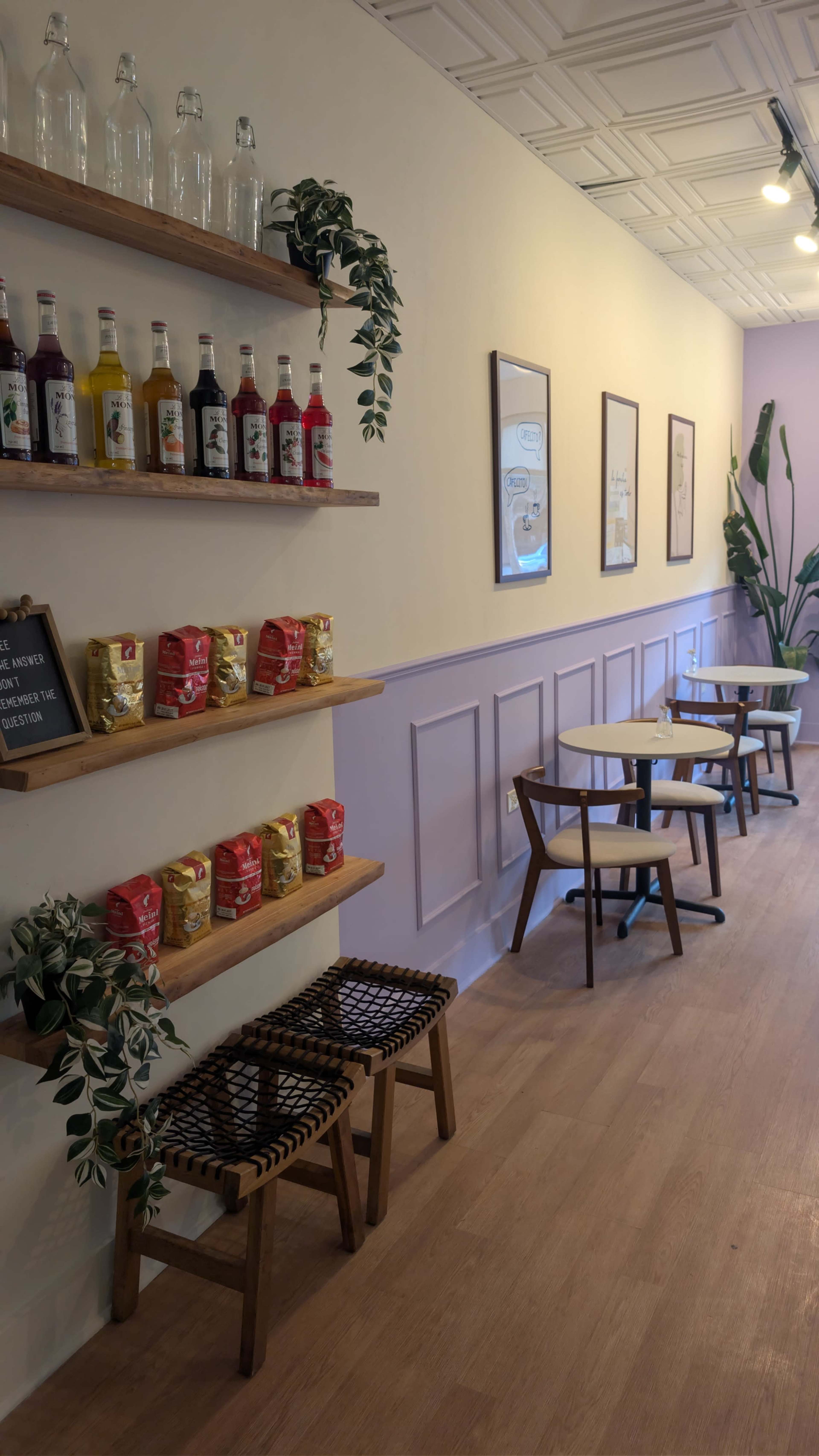 The image shows a cozy café interior with wooden shelves displaying drinks and snacks, and small tables set up against a light-colored wall.