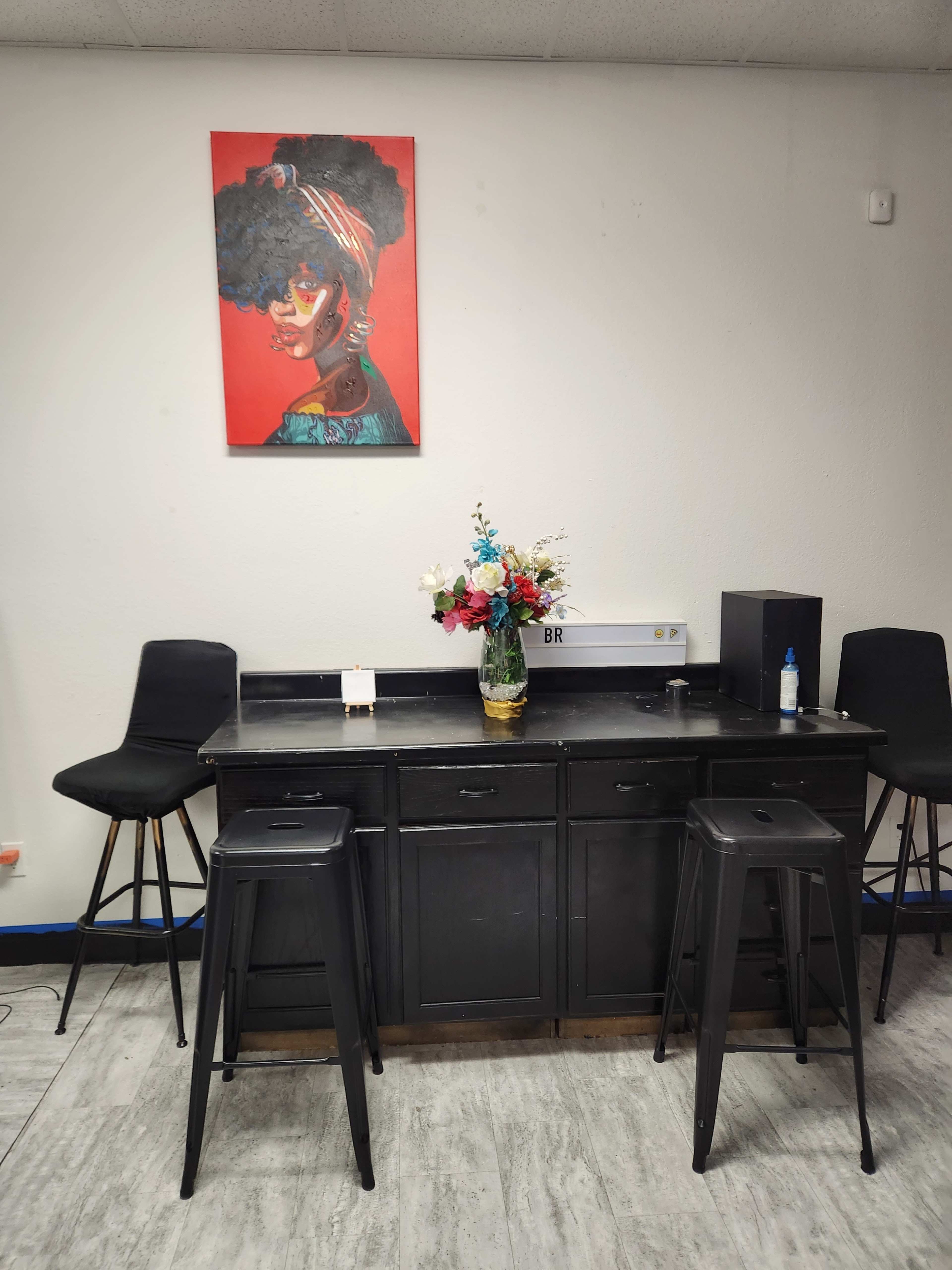 A black countertop with two metal stools, a vase of flowers, and a colorful portrait hanging on the wall above.