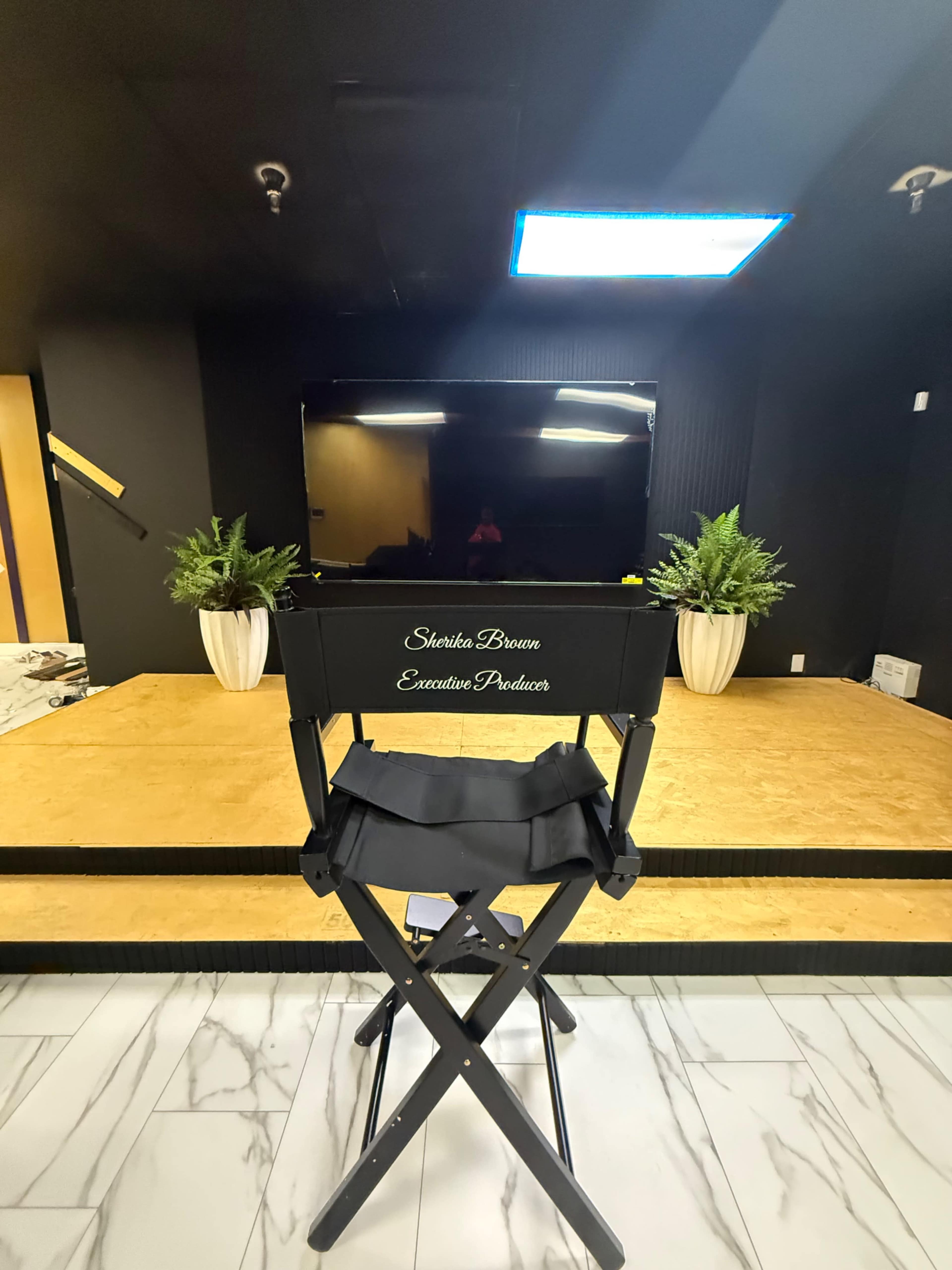 A director's chair with the name "Shanta Brown" and the title "Executive Producer" is positioned facing a black television set on a wooden stage with potted plants on either side.