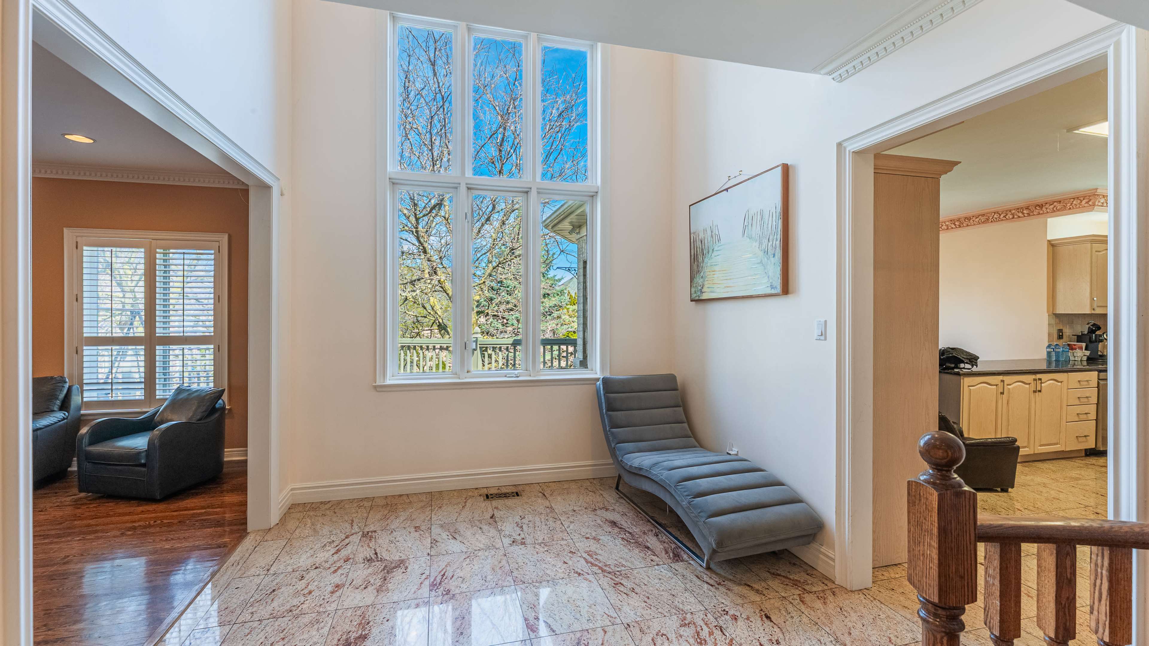 A spacious foyer features a sleek chaise lounge beside large windows illuminating the area, with a view of trees outside.