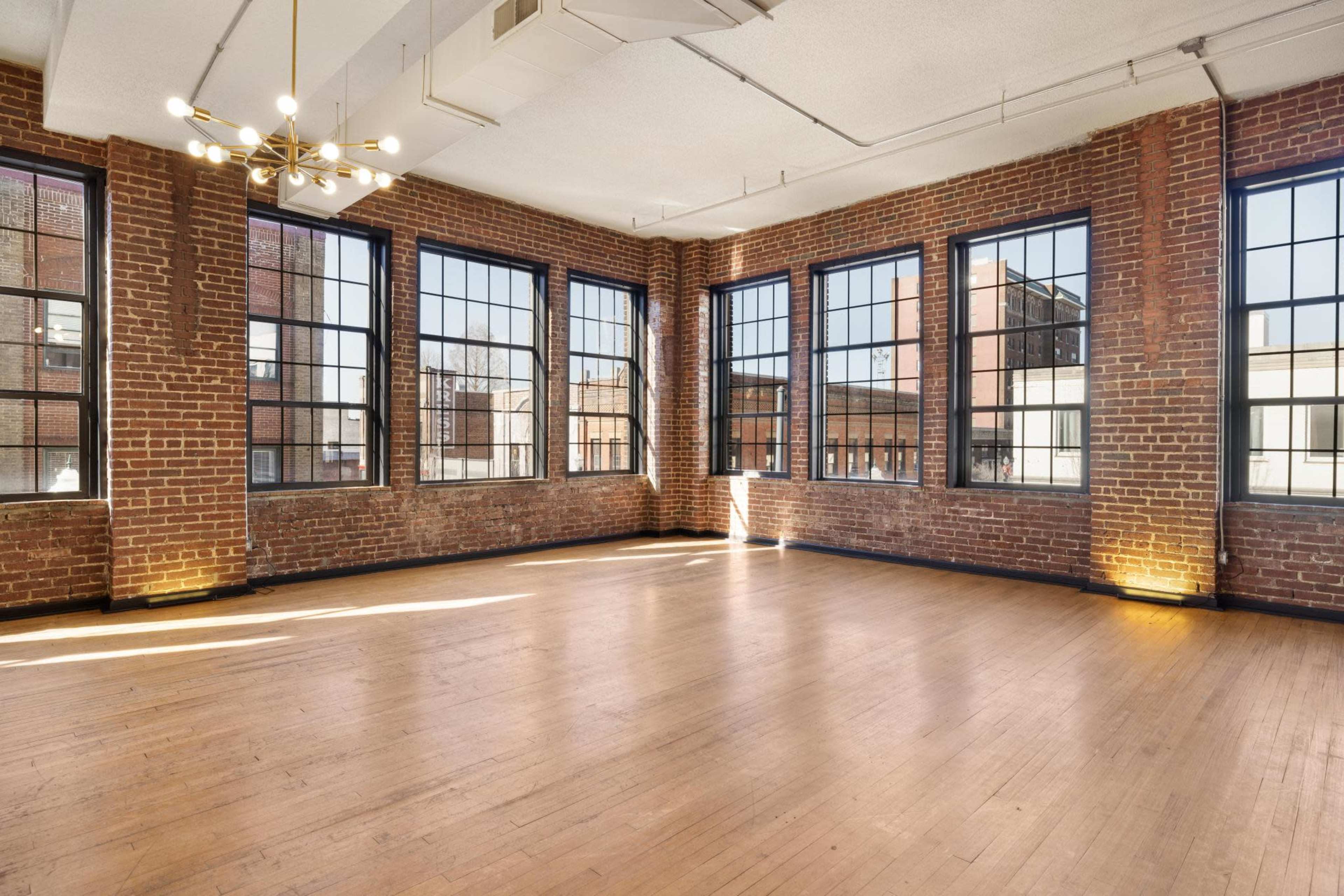 A spacious room with exposed brick walls, large windows, and wooden flooring.