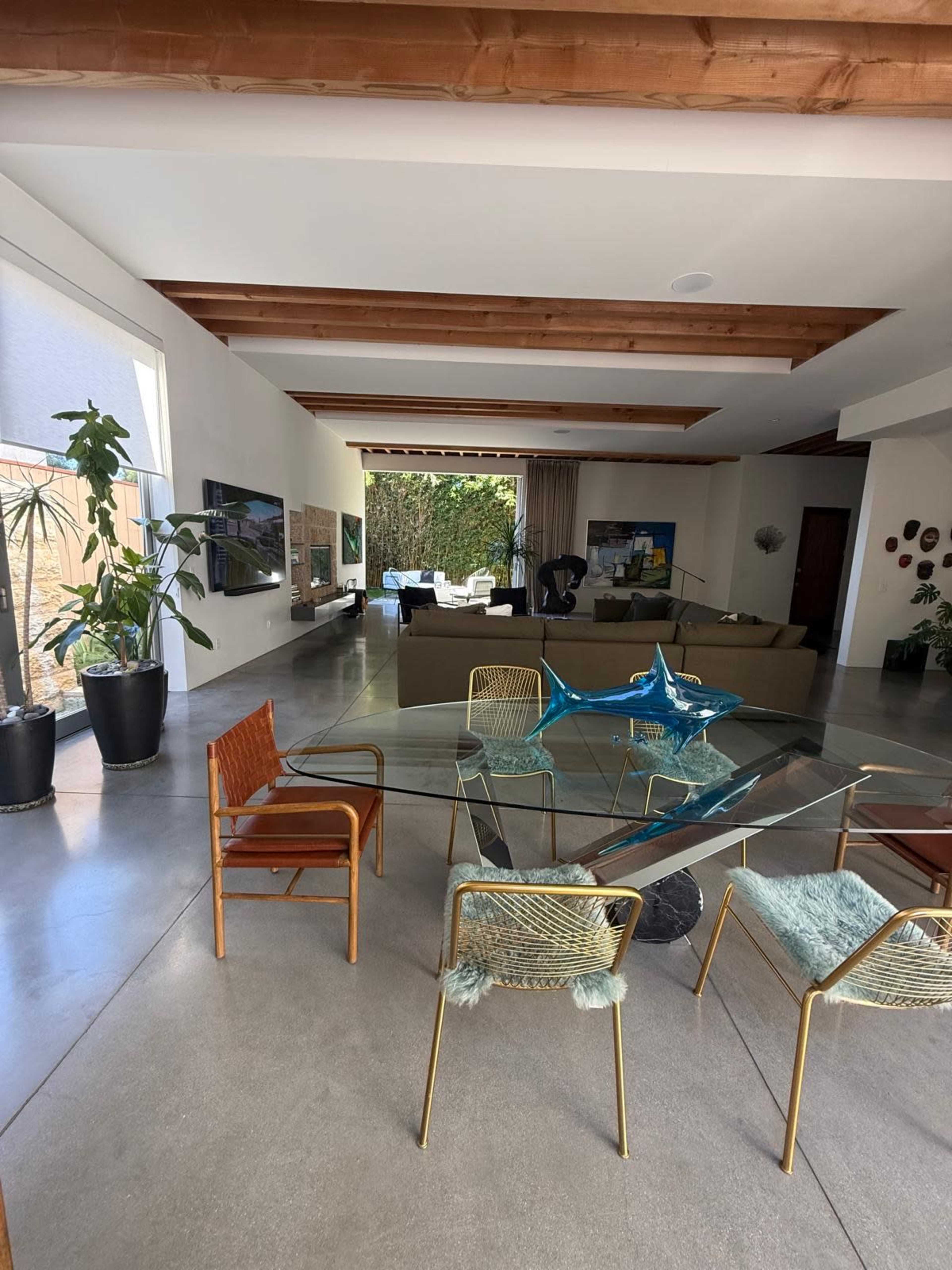 The image shows a modern, open-plan living space featuring a glass dining table surrounded by various chairs, with a large sectional sofa and a television in the background.