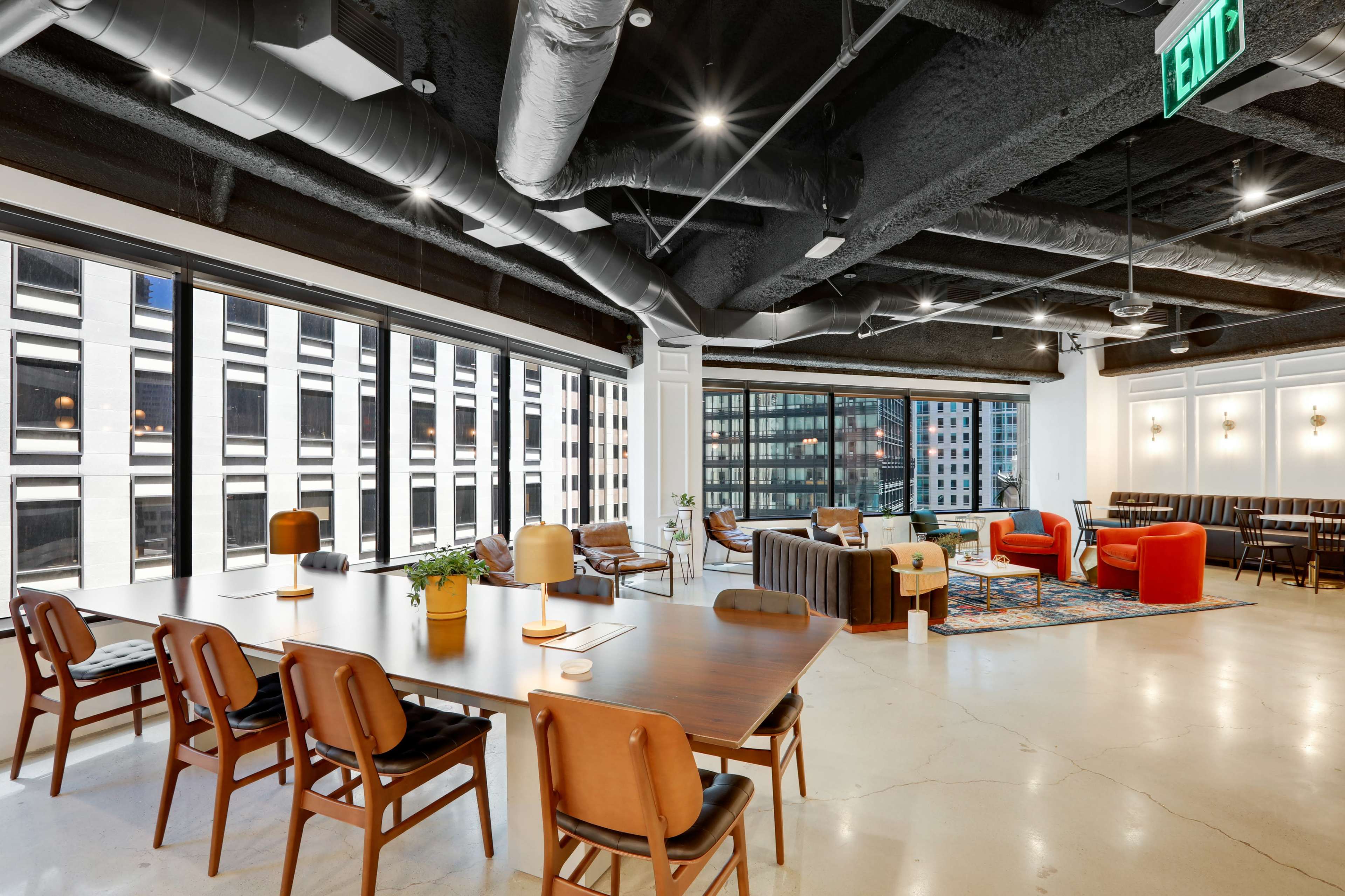 A spacious modern office interior with large windows, a mix of seating areas, and a dining table surrounded by chairs.