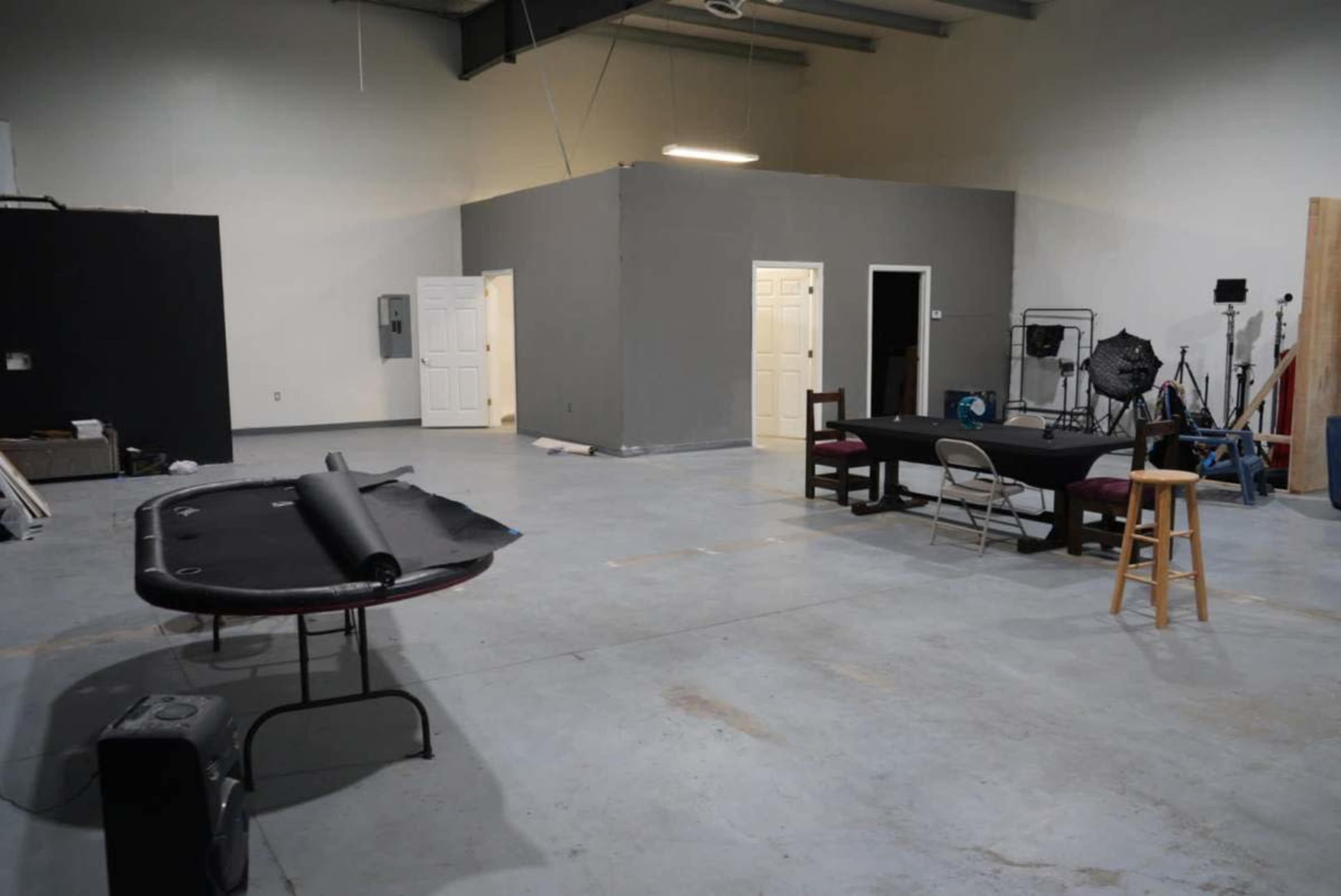 The image shows an empty industrial studio space with a gray wall, a table with chairs, and a roll of black material on a table.