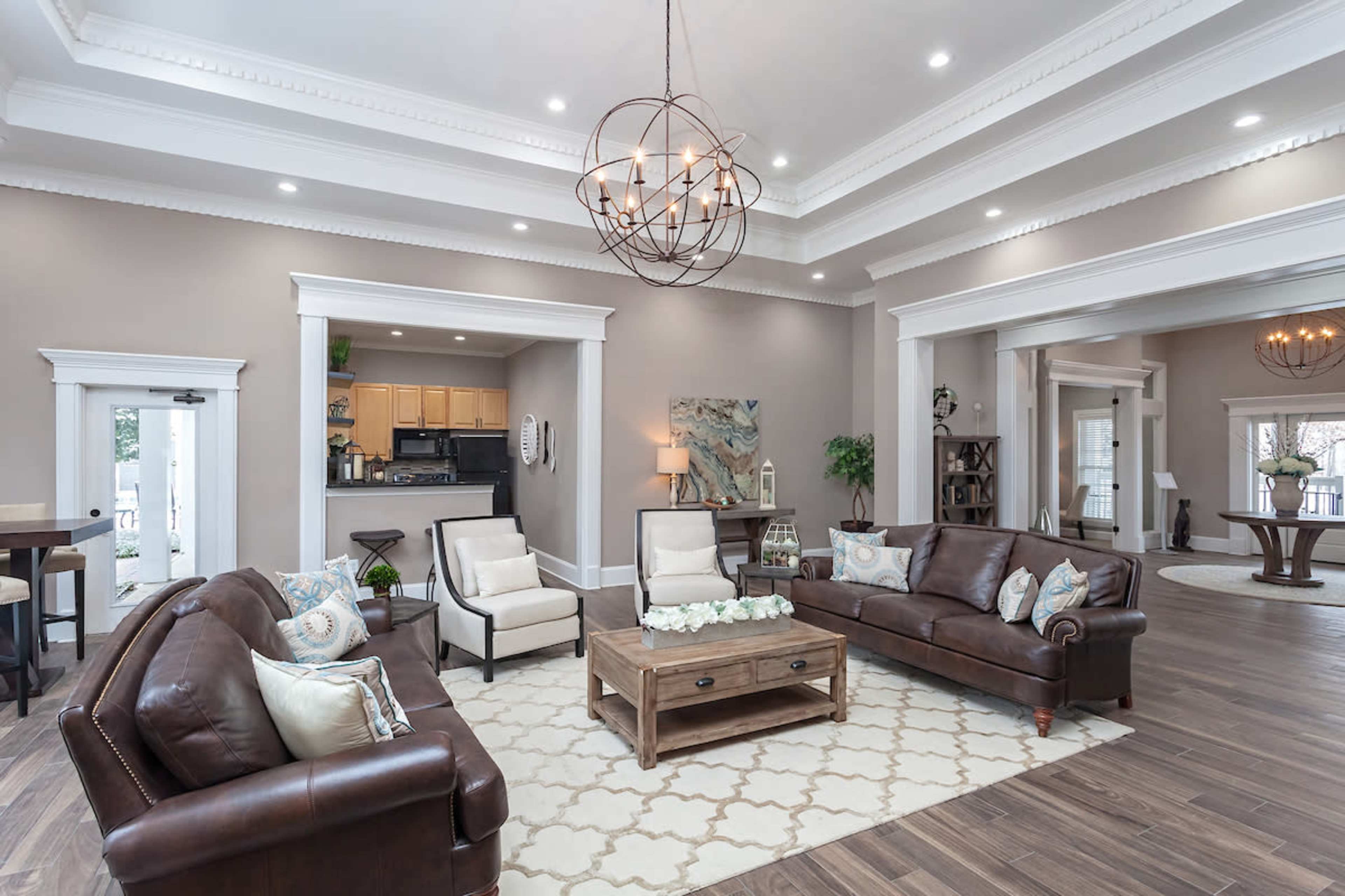 A spacious living room featuring a combination of brown leather sofas, a light-colored area rug, a wooden coffee table, and a modern chandelier, with an open view towards a kitchen area.