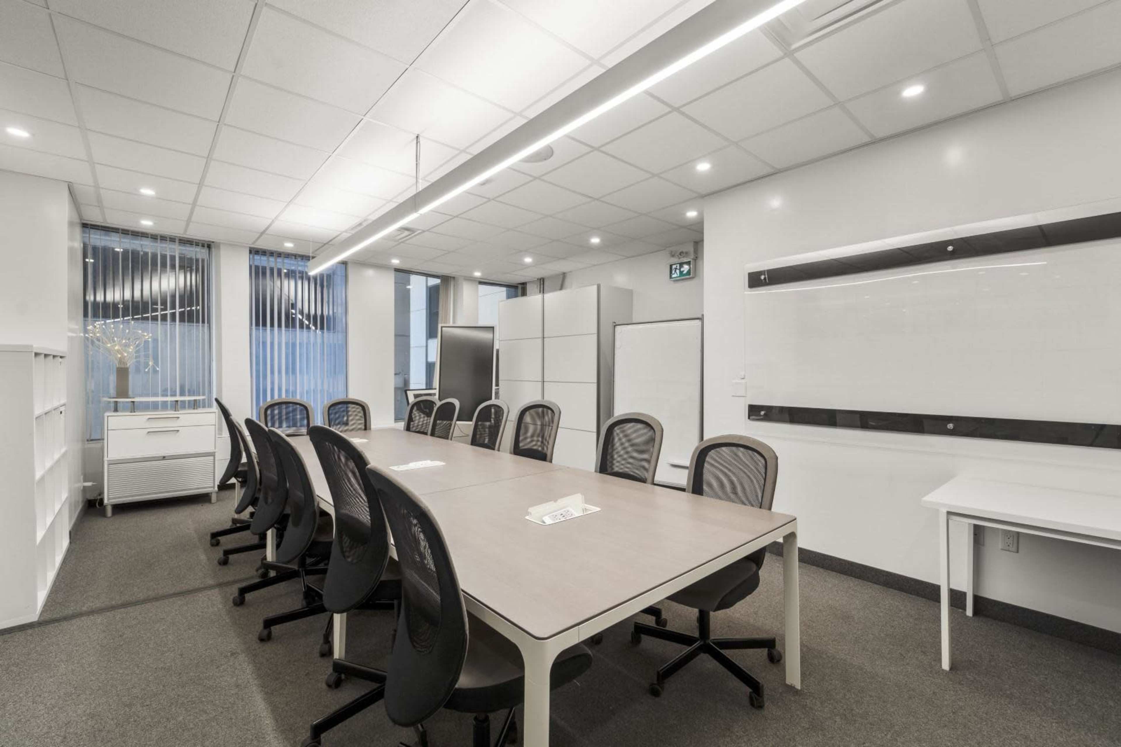 A modern conference room with a long table surrounded by black office chairs, large windows, and a whiteboard on the wall.