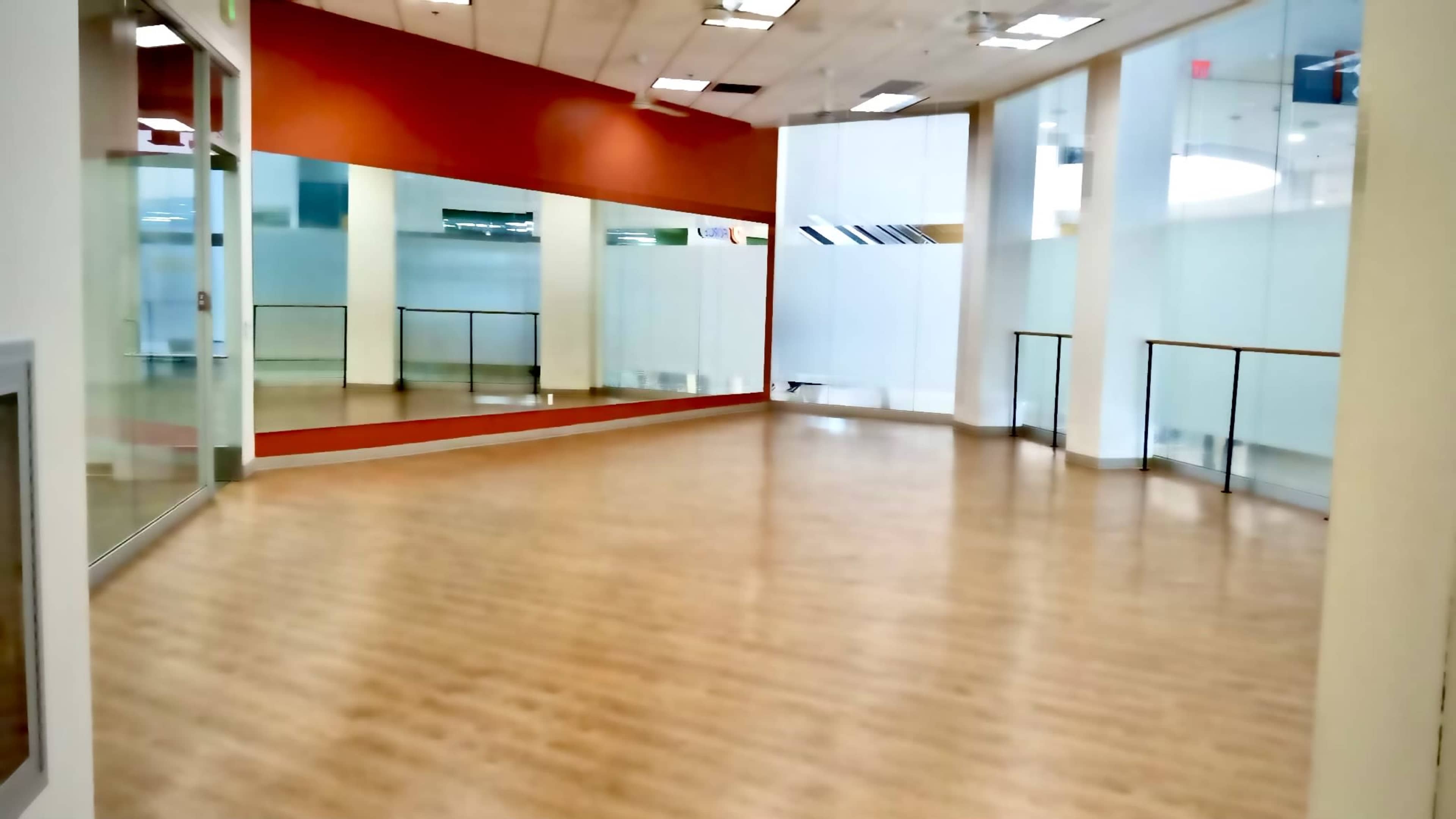 The image shows a spacious, empty room with wooden floors and large mirrors on one wall.