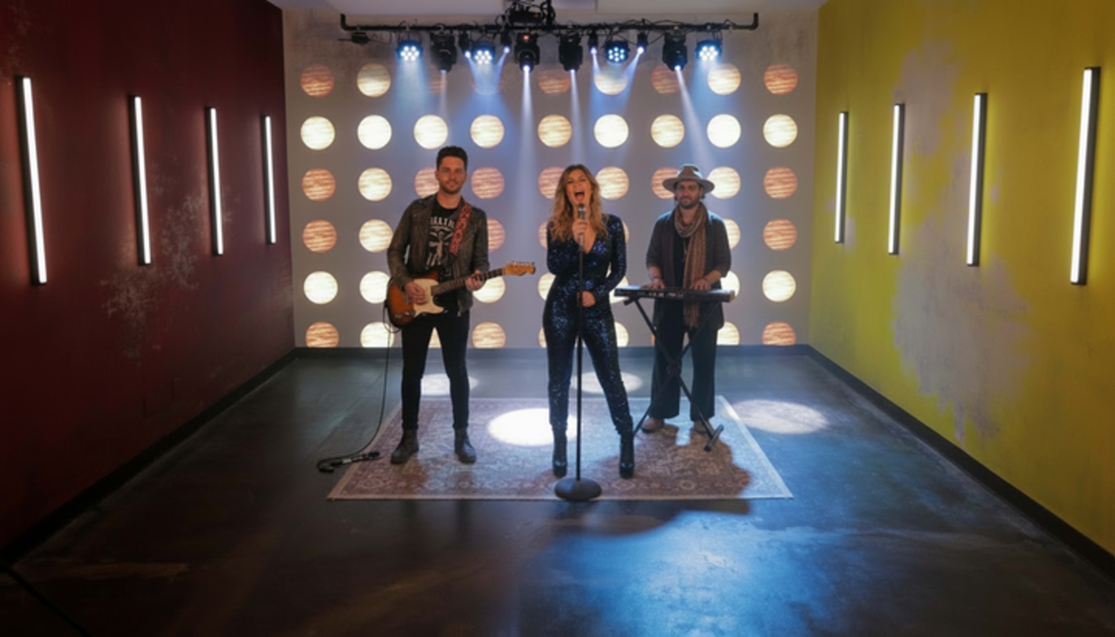 A music group stands performing on a stage with colorful lights and a patterned backdrop.
