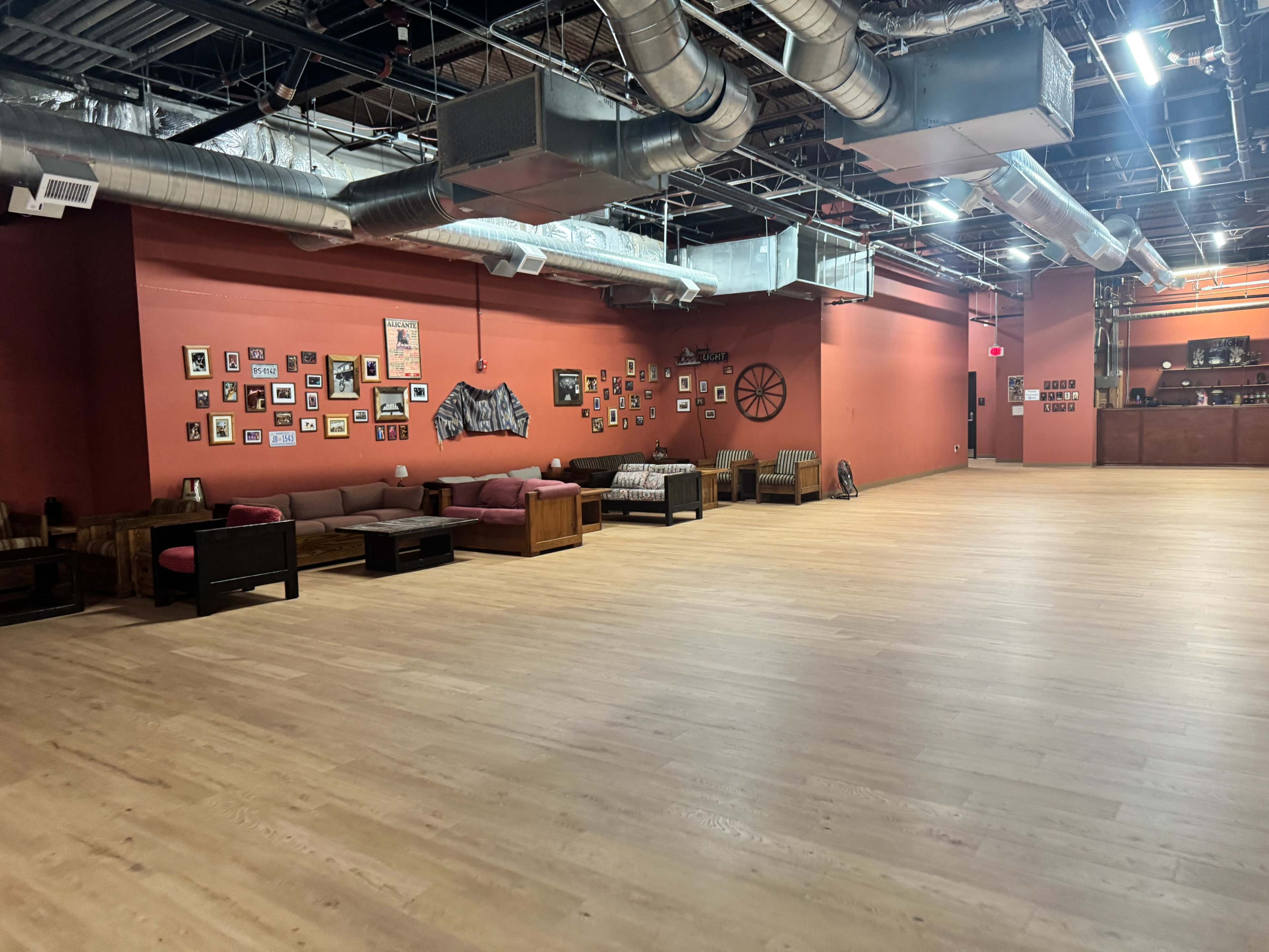 The image shows a spacious room with wooden flooring, a red-painted wall adorned with various framed photographs, and seating arrangements including sofas and chairs.