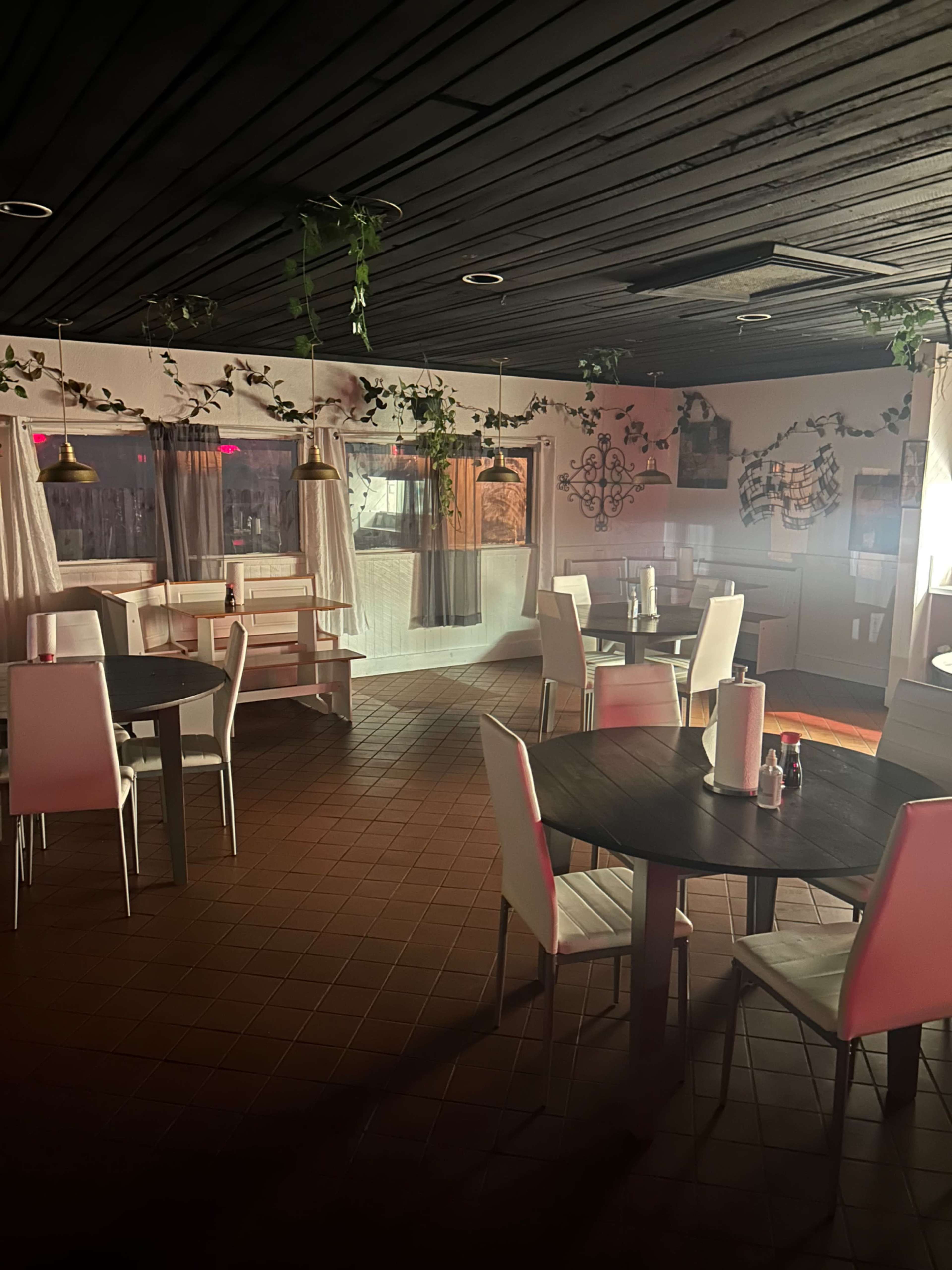 The interior of a restaurant features several round tables with white chairs, surrounded by indoor plants and soft lighting.