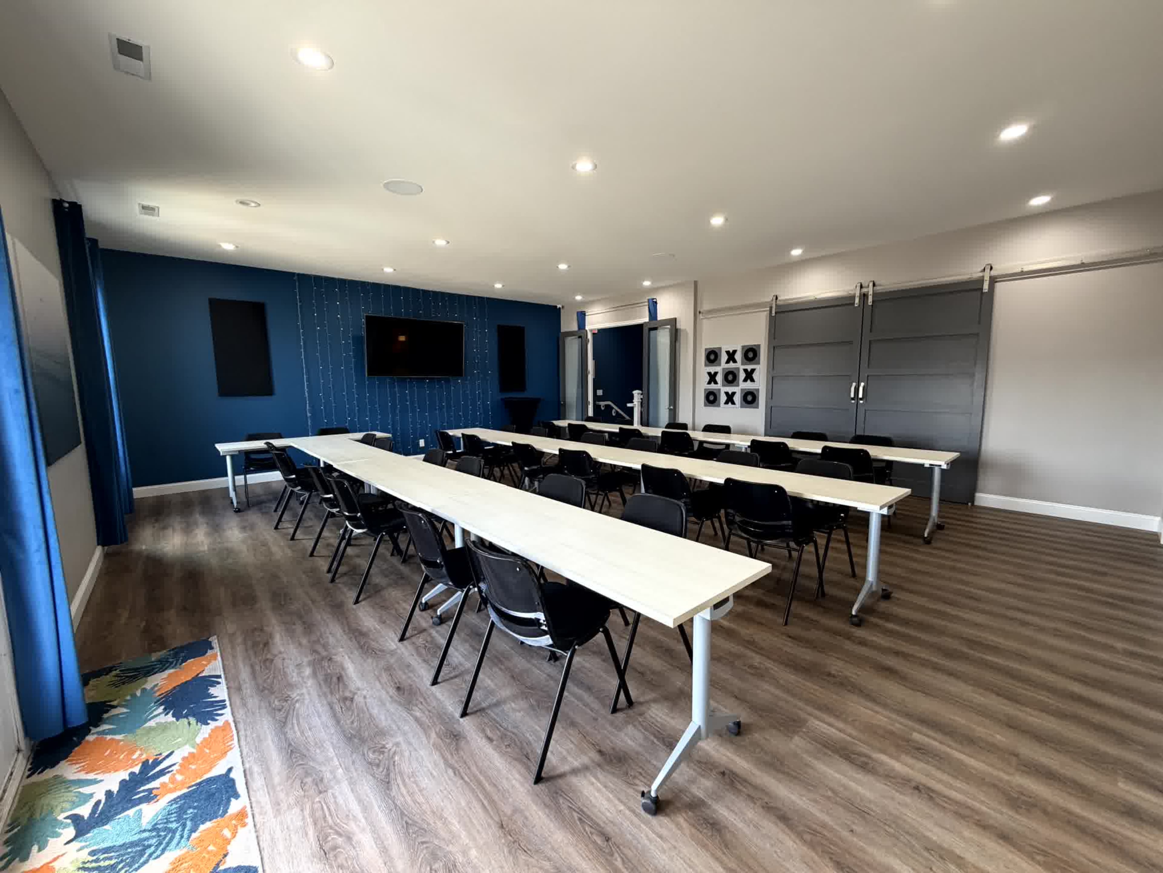 The image shows a conference room featuring several rows of long tables and black chairs, with a blue accent wall and a large screen on one side.