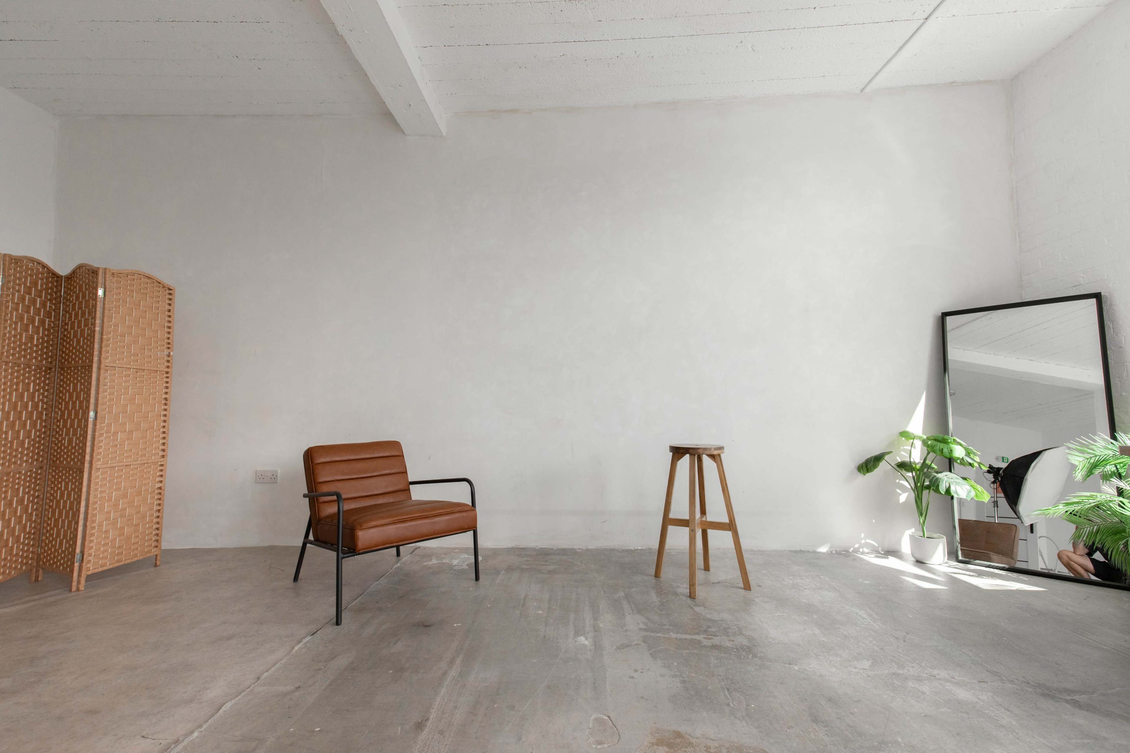 A minimalist interior space with a brown leather chair, a wooden stool, a tall mirror, and a potted plant against a light-colored wall.