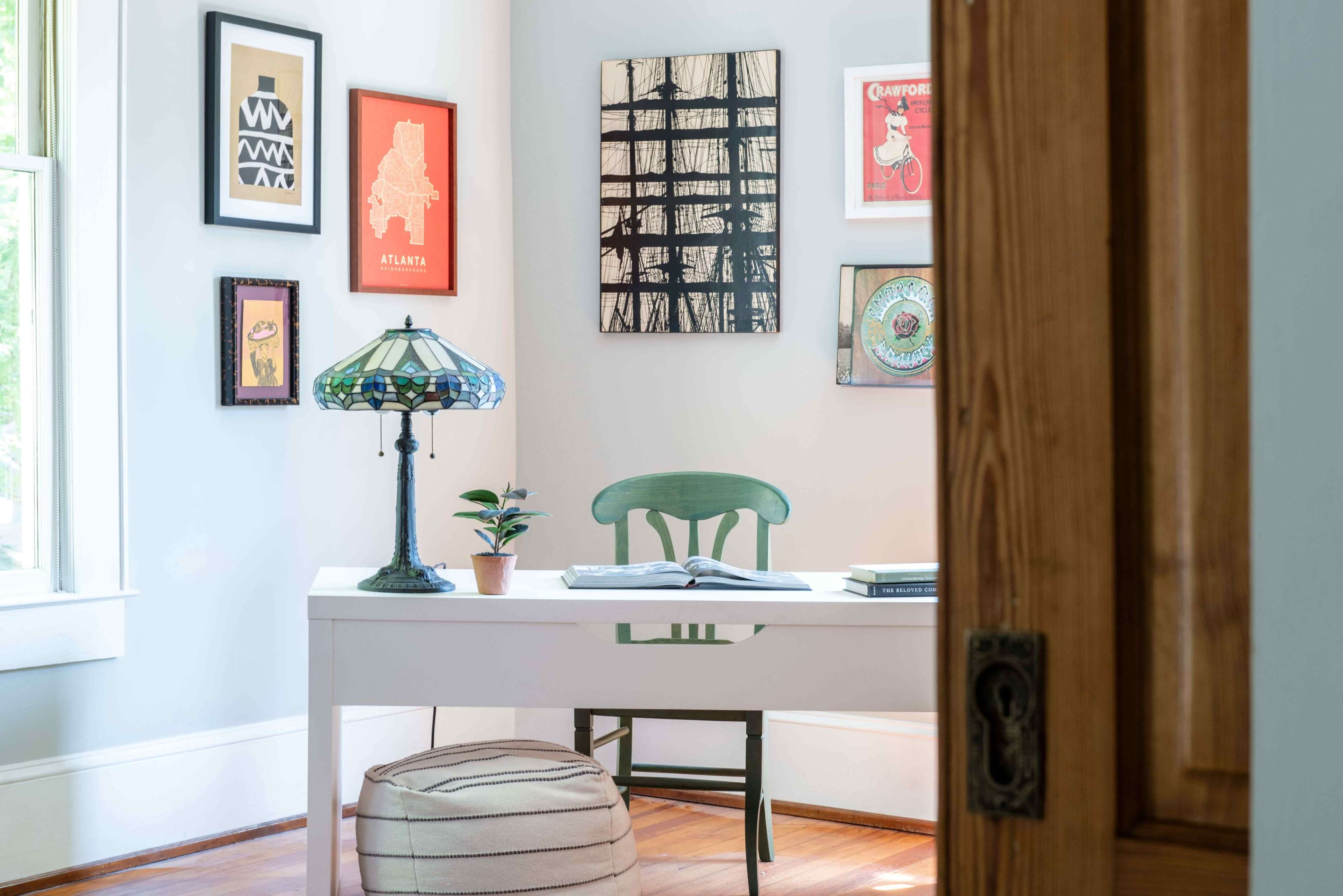 A small workspace features a white desk with an open book, a green chair, a potted plant, and a stained glass lamp, surrounded by framed artwork on the walls.