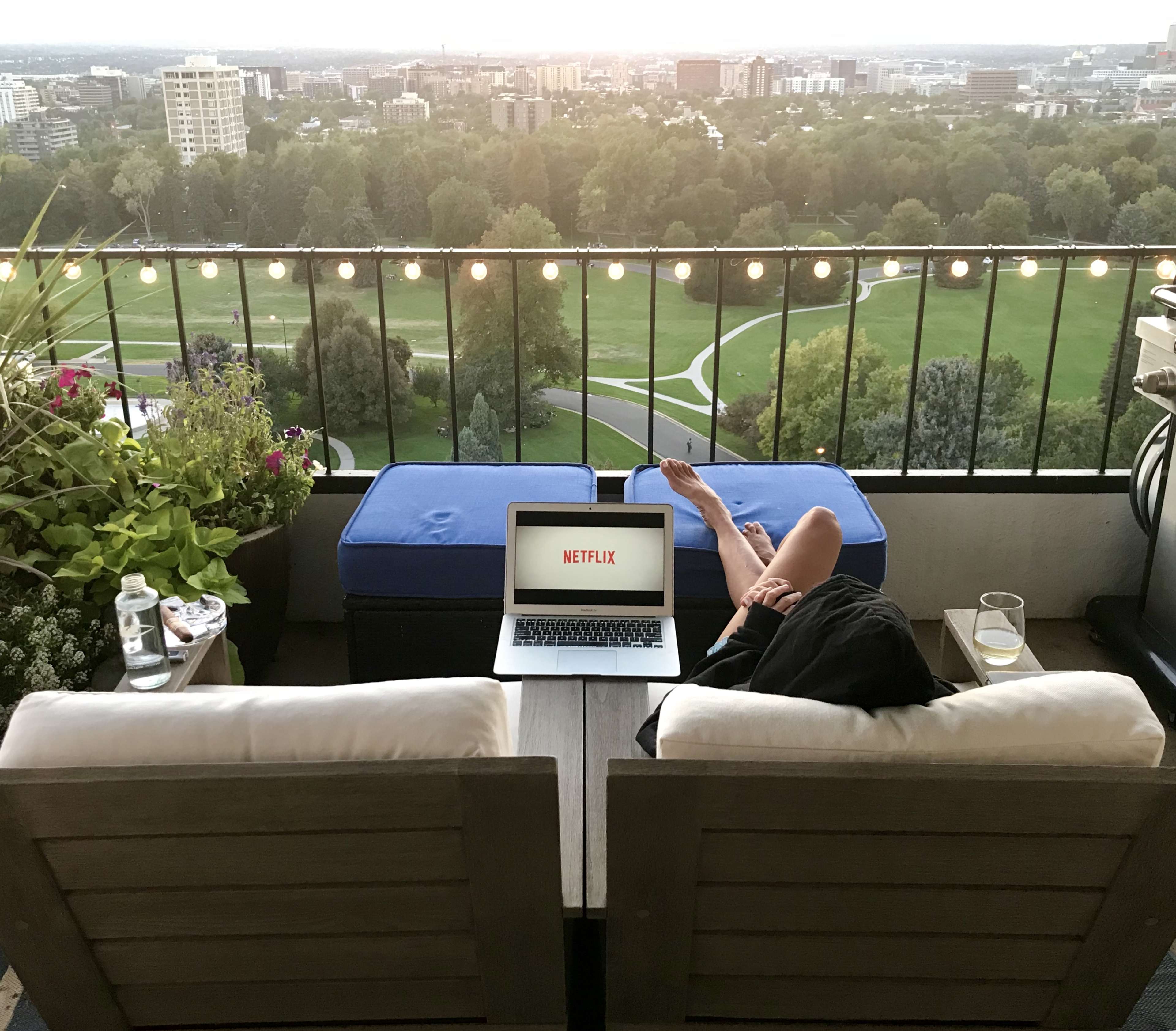 A person relaxes on a balcony with a laptop displaying Netflix, overlooking a city skyline at sunset.