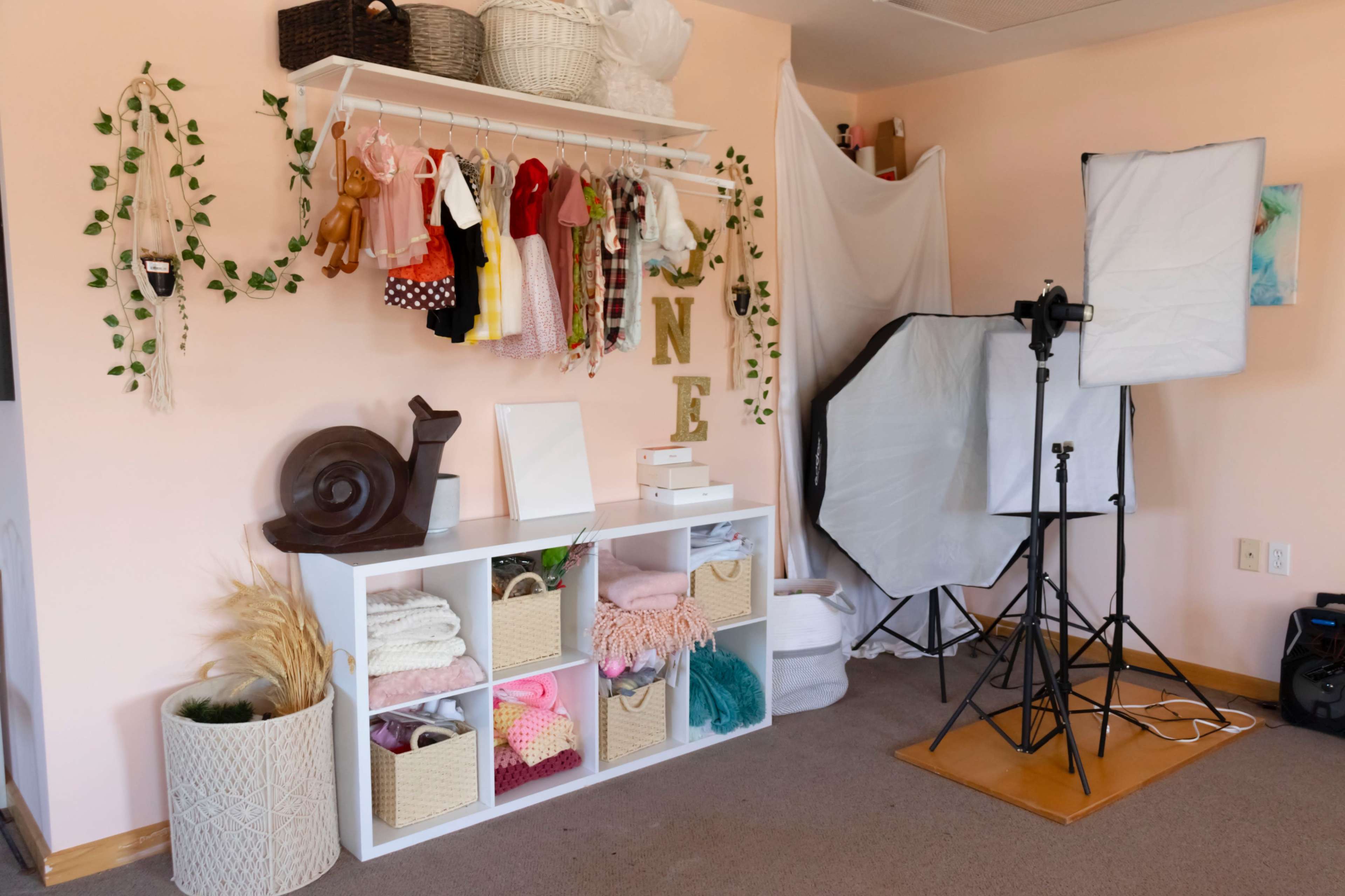 A room with a shelf displaying various children's clothing, a white storage unit filled with folded fabrics and accessories, and photography equipment set up in the corner.