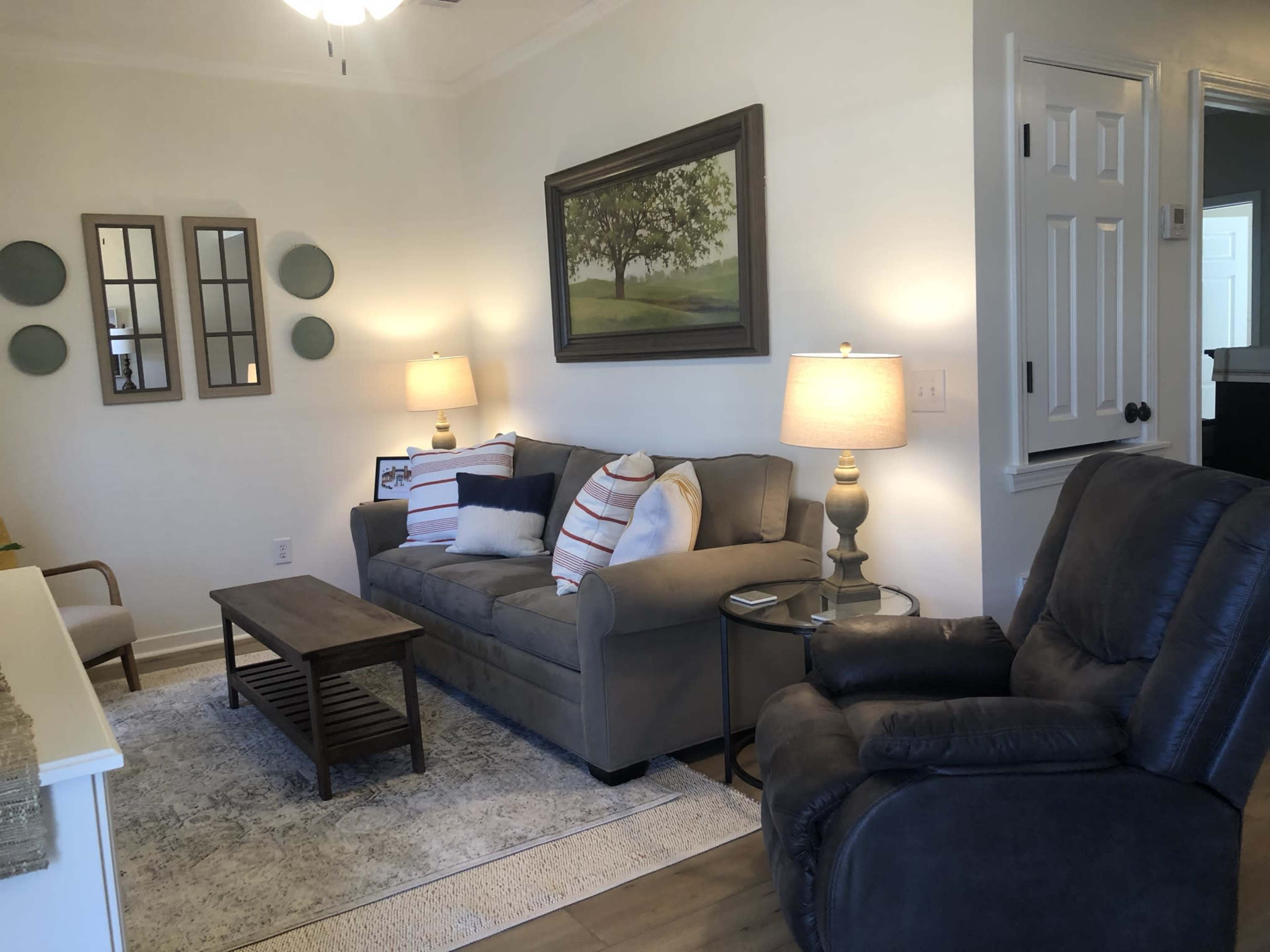 A living room with a gray sofa, a coffee table, two lamps, and an armchair, complemented by wall decor featuring mirrors and a landscape painting.