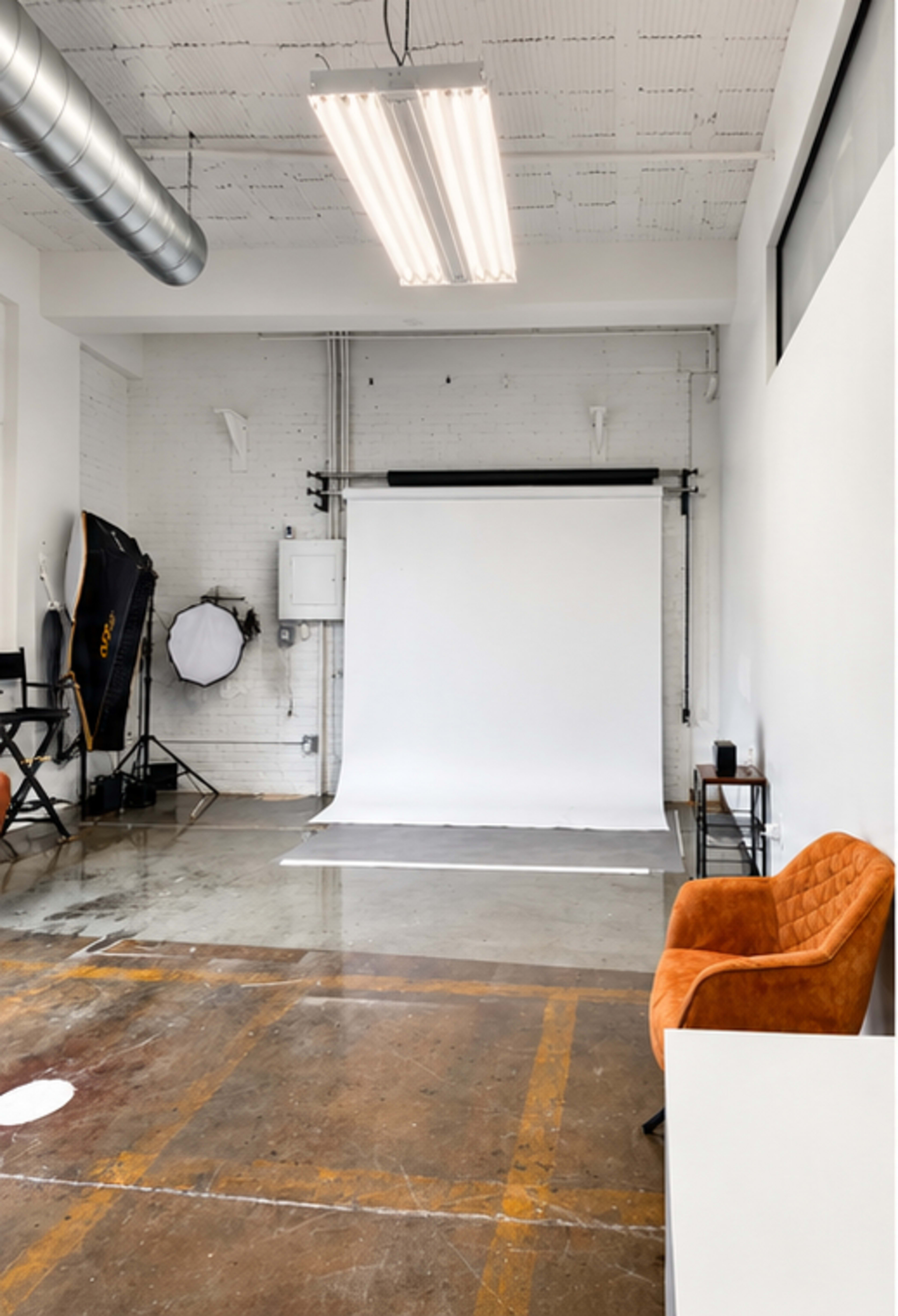 The image shows a photography studio with a white backdrop, studio equipment, and an orange chair in the corner.
