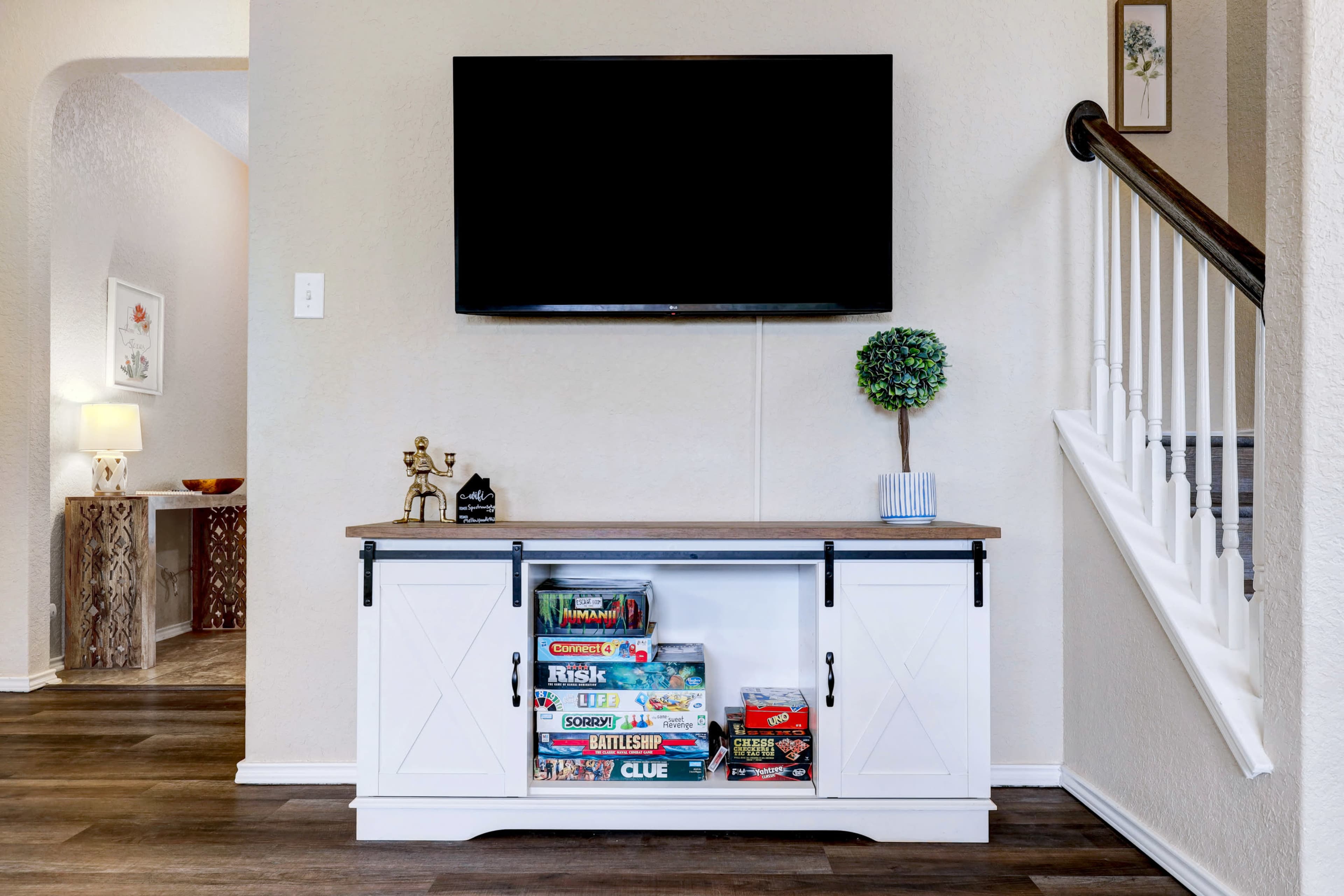 A white TV stand with board games displayed inside sits beneath a wall-mounted television in a well-lit room.