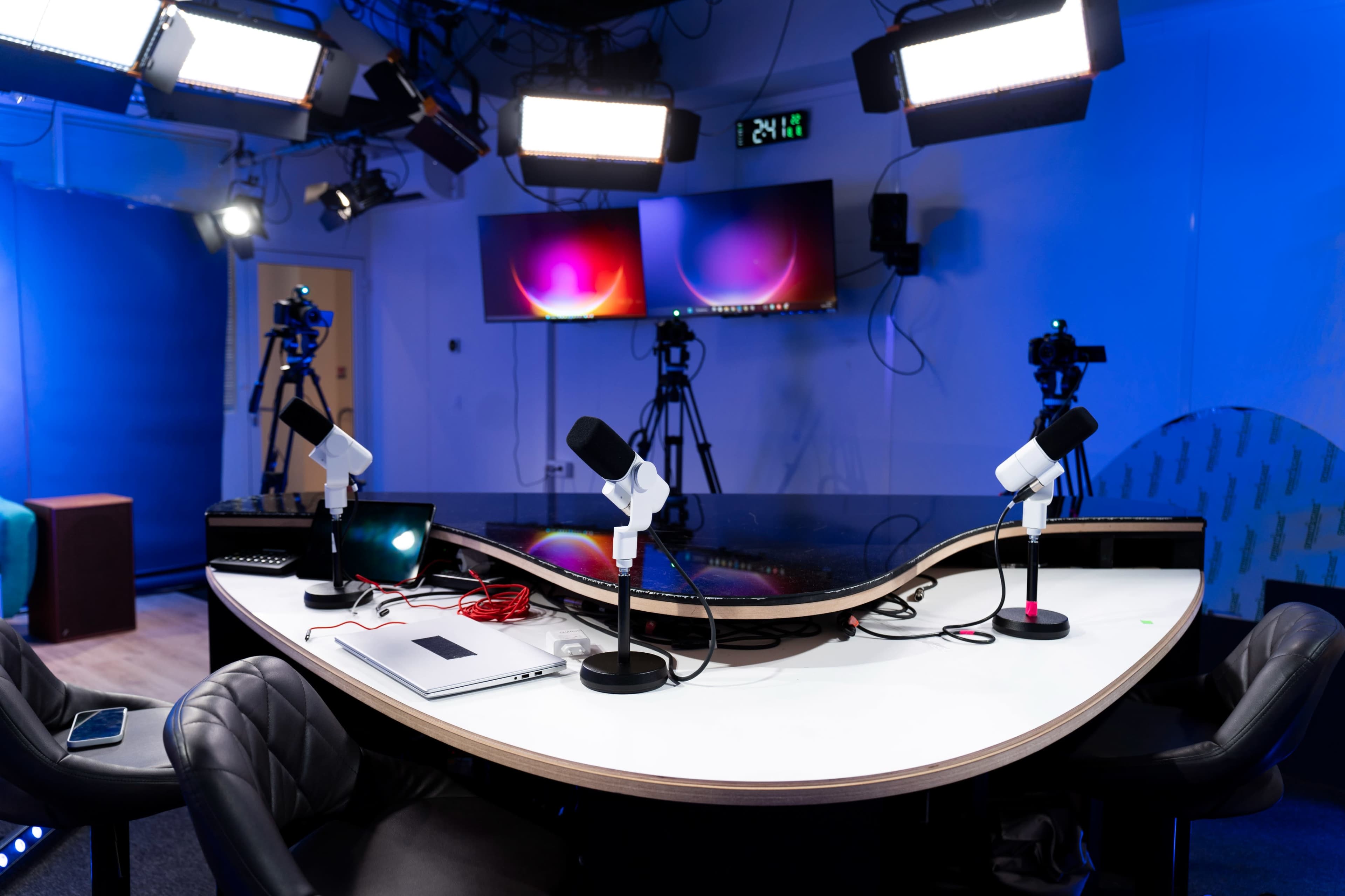 A modern studio setup features a curved table with microphones, laptops, and multiple cameras, illuminated by overhead lights against a blue backdrop.