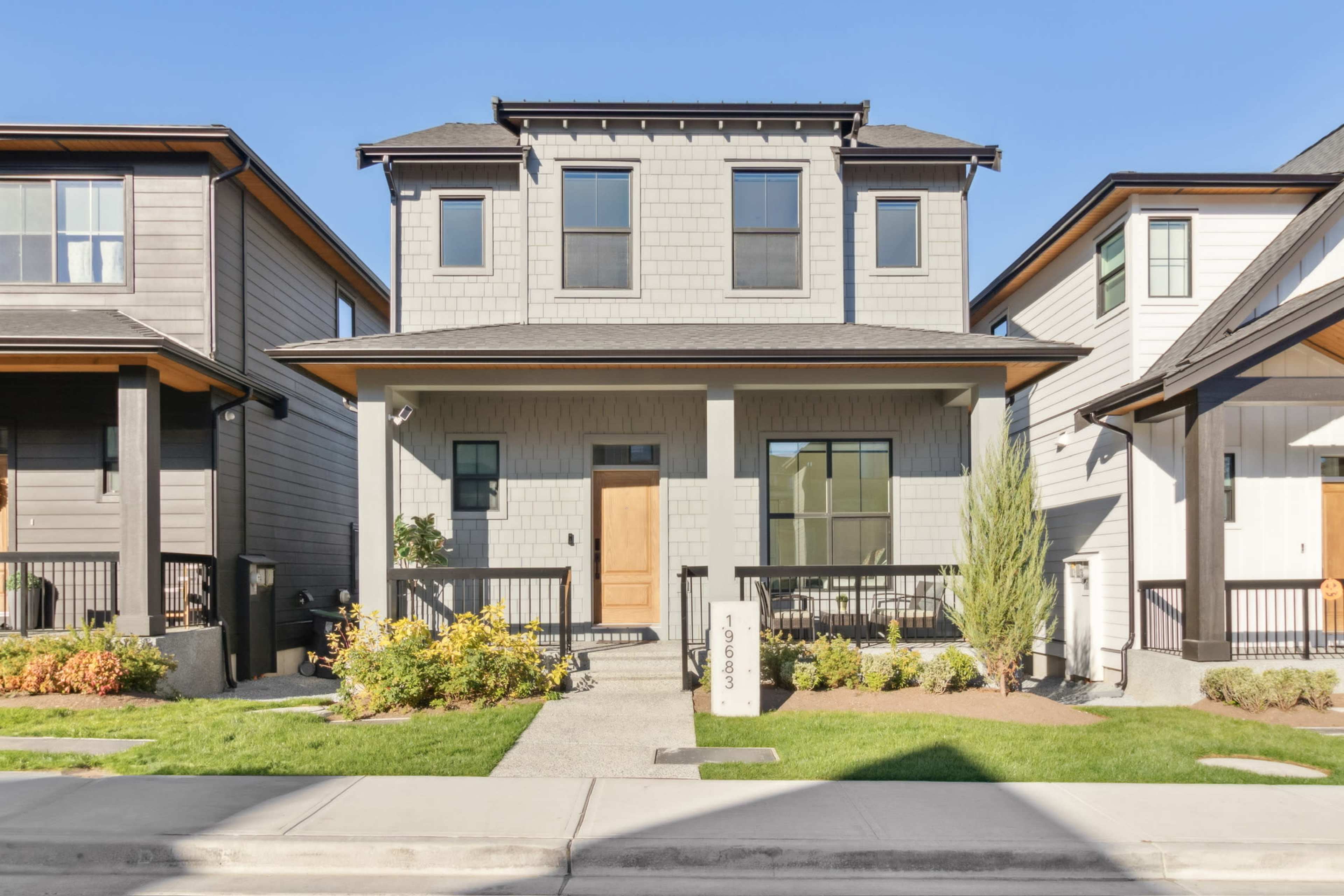 The image shows a two-story gray house with a wooden front door and landscaped yard, flanked by two similar homes on either side.