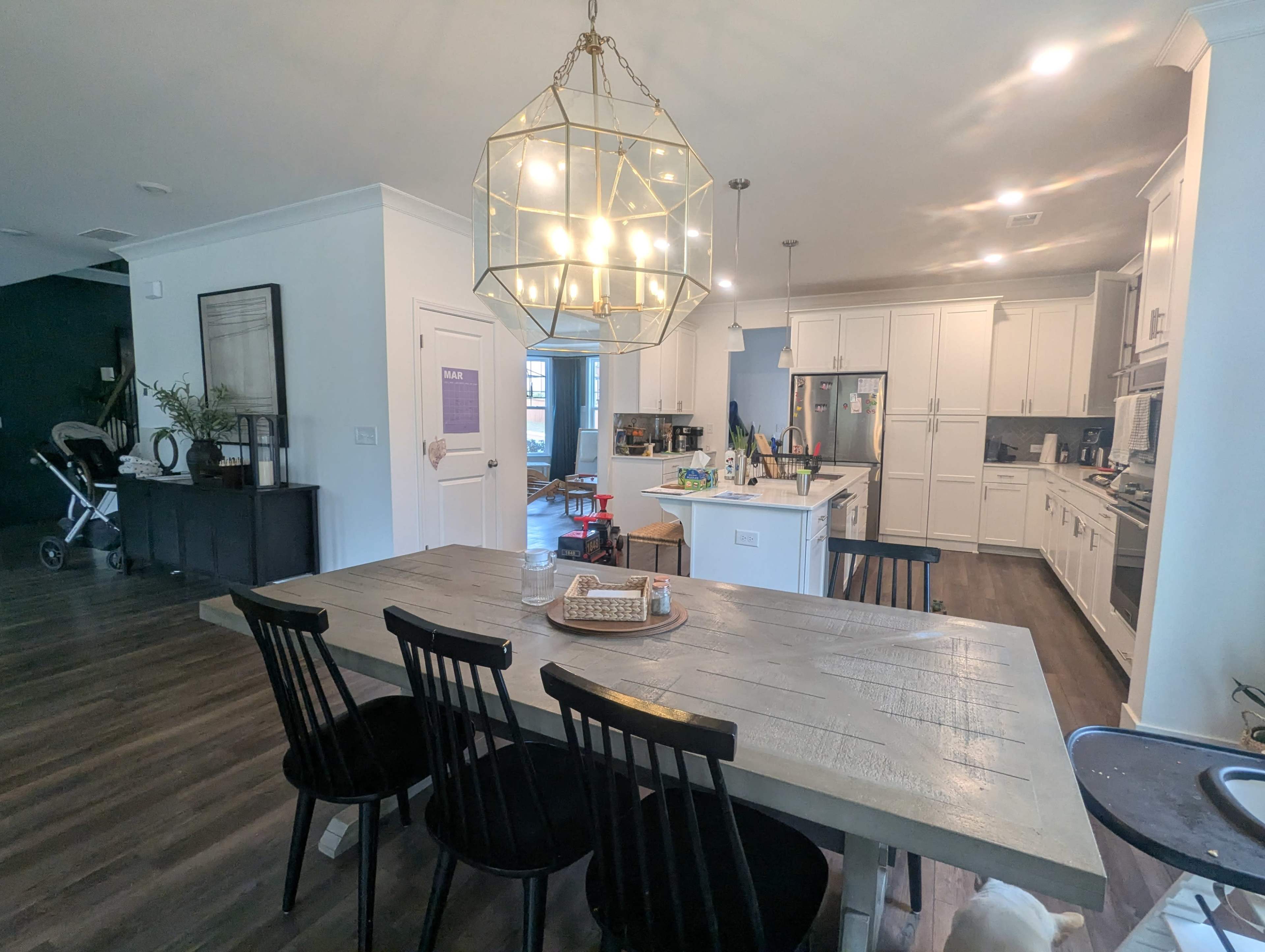 A spacious kitchen and dining area features a large wooden table with black chairs, a modern chandelier, and white cabinetry with stainless steel appliances.