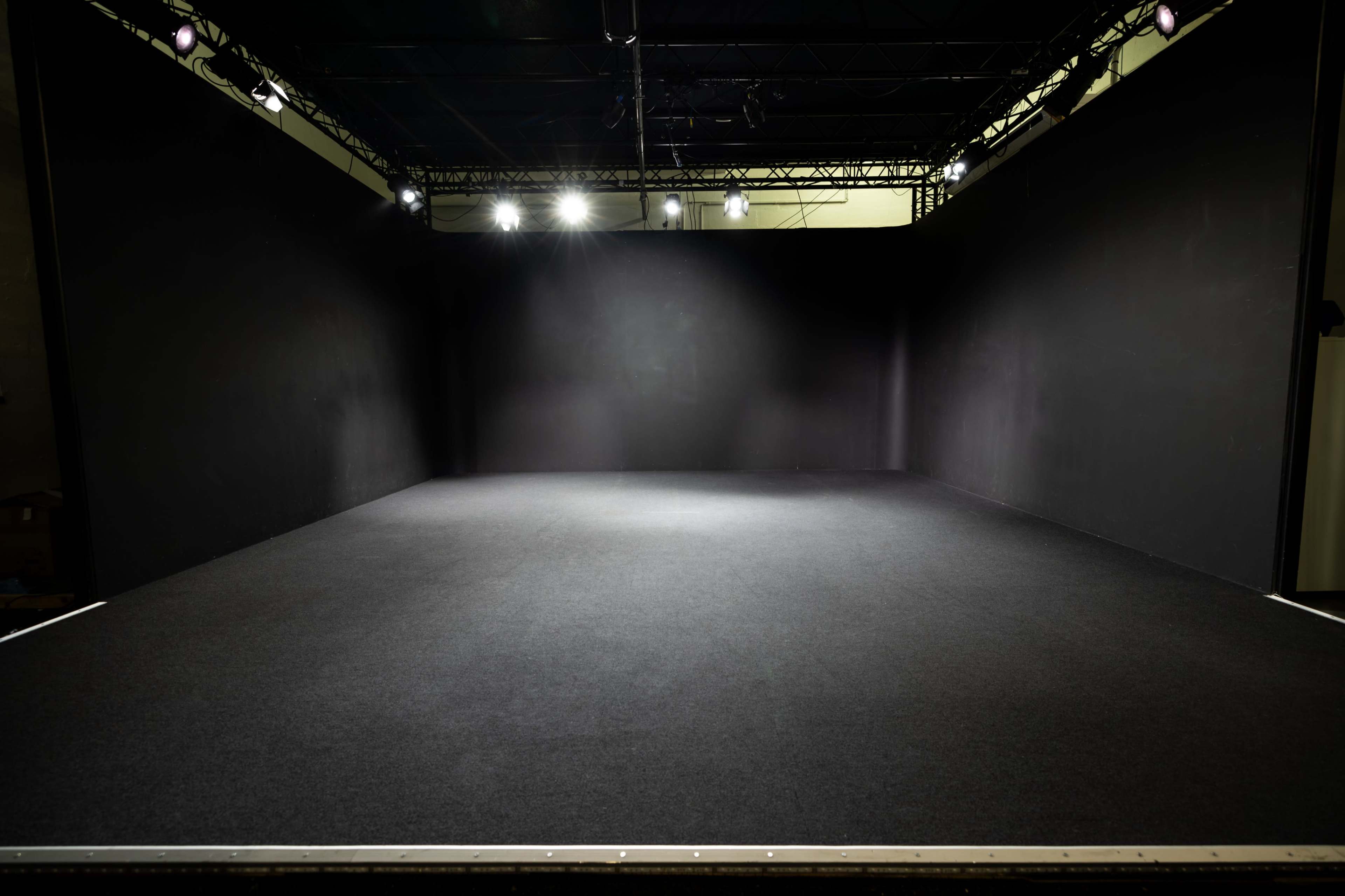 A large empty stage with black walls and a dark carpeted floor, illuminated by overhead lighting.