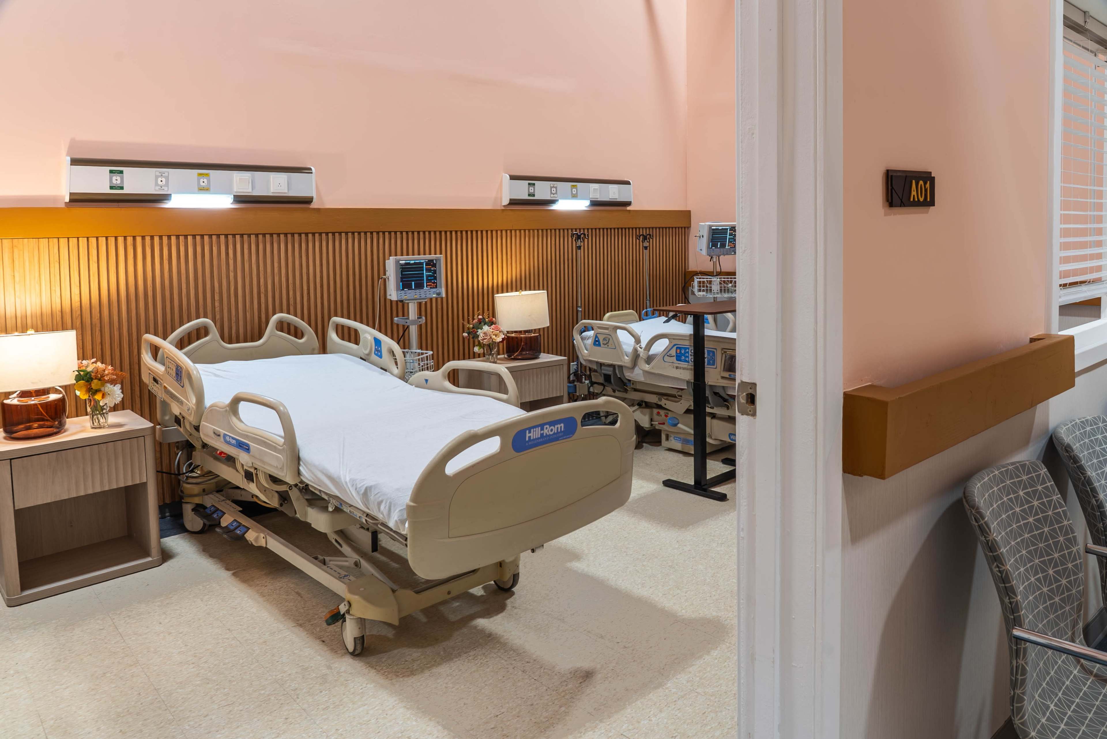 The image shows a hospital room with two empty patient beds, each equipped with medical monitors and bedside tables, adjacent to a doorway marked "A01."