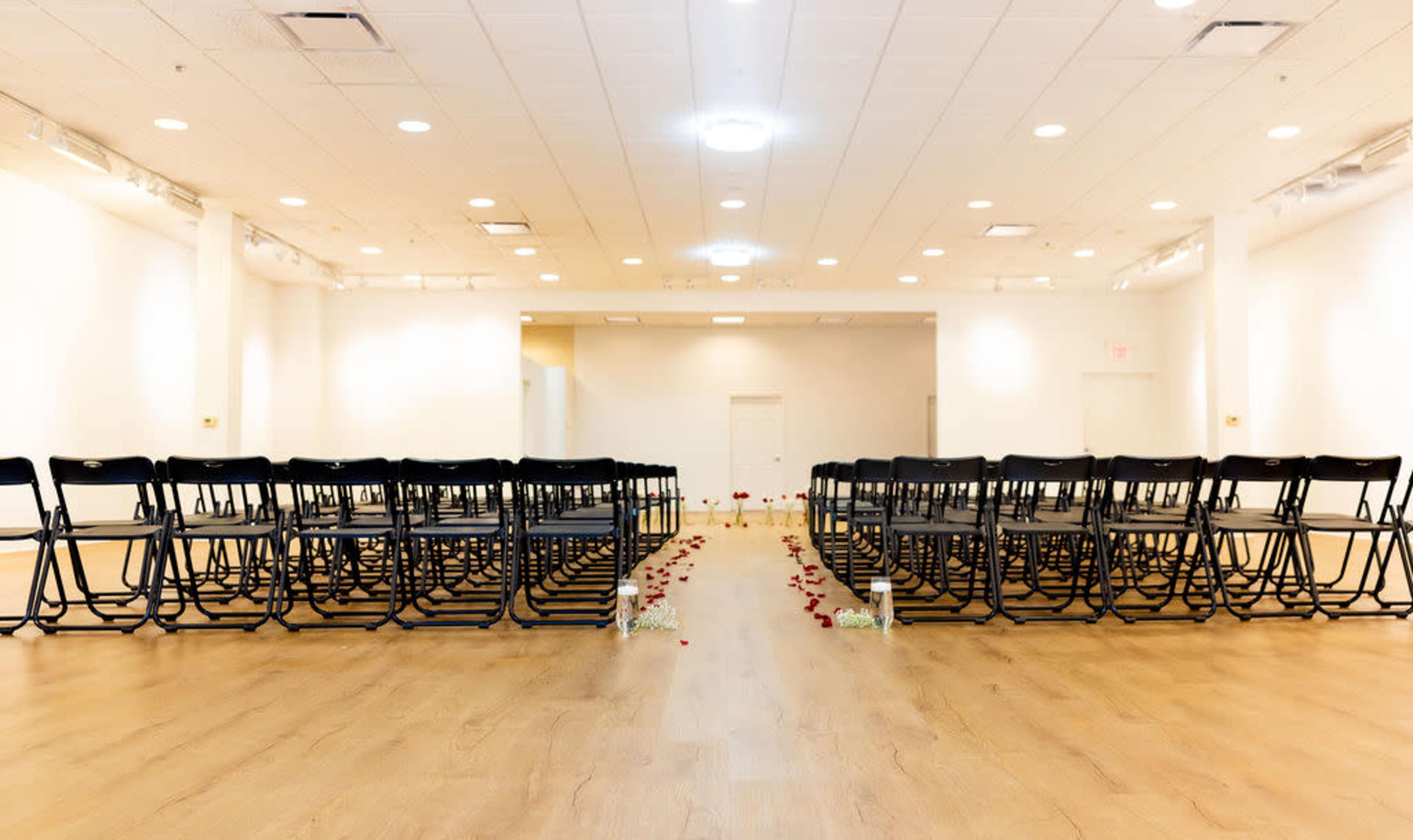 The image shows a spacious indoor venue set up for a ceremony, with rows of black chairs arranged facing a center aisle adorned with rose petals.