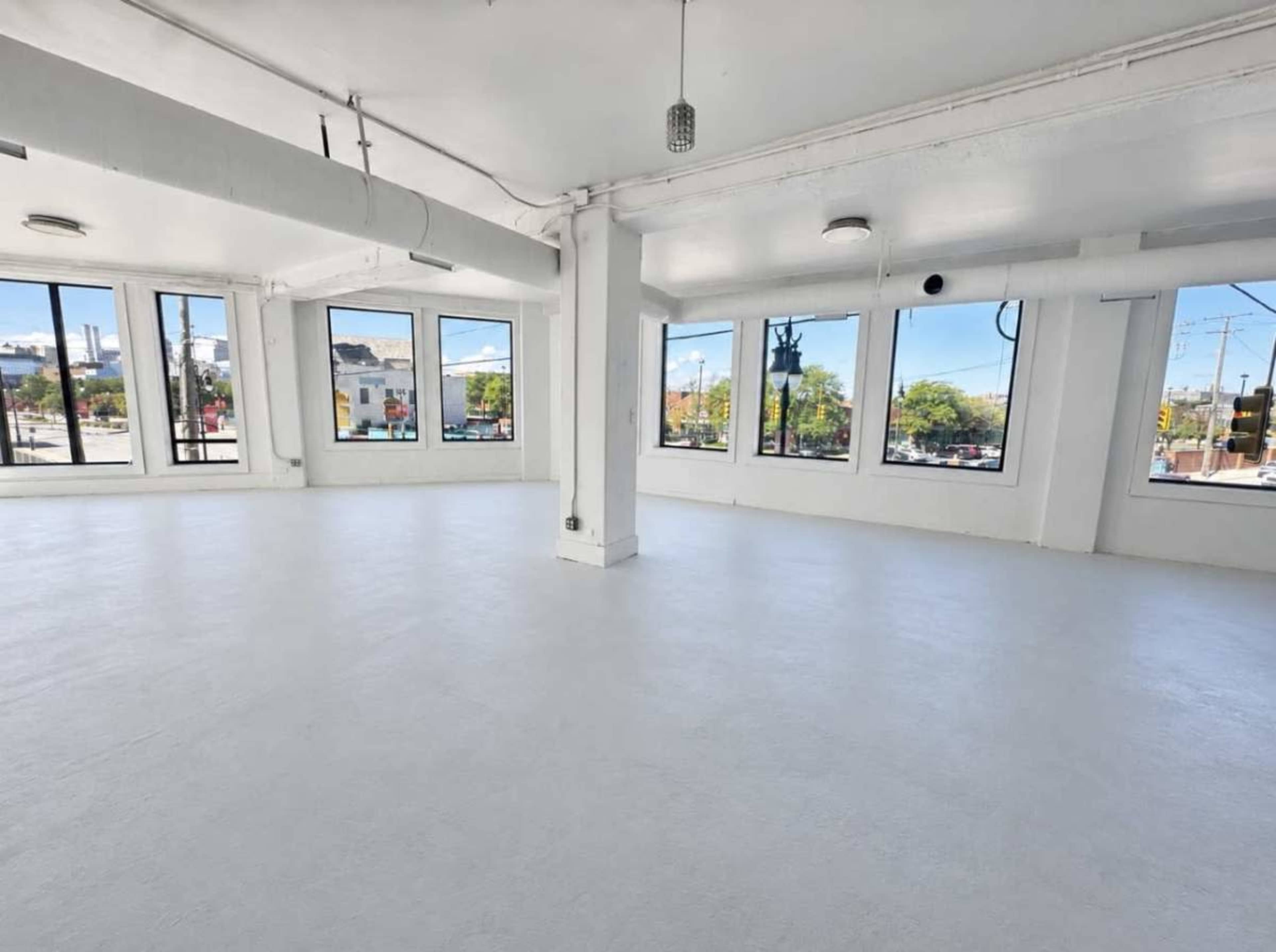 The image shows an empty room with white walls and a light-colored floor, featuring large windows that let in natural light.