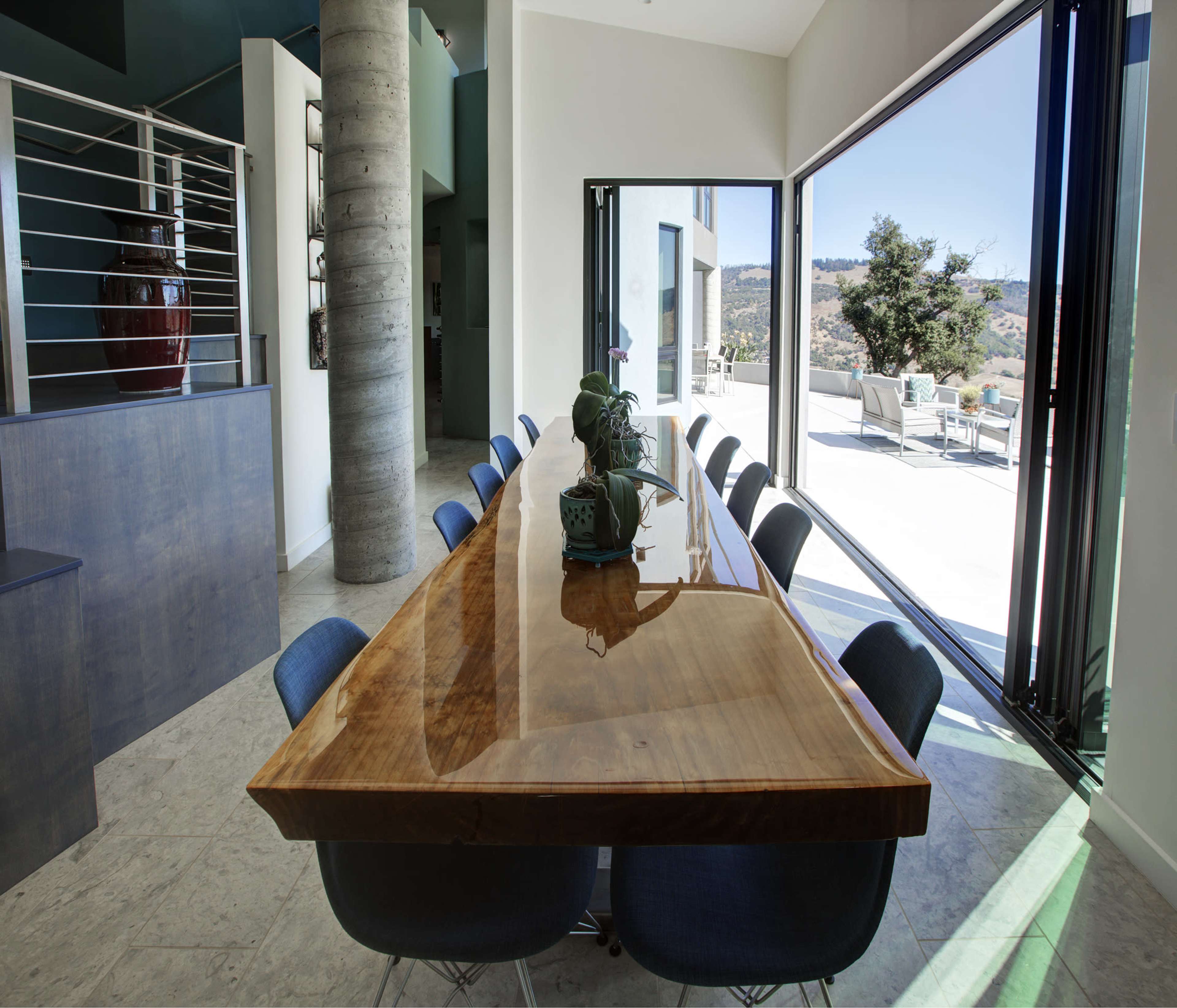A long wooden dining table with blue chairs is positioned in a modern interior space featuring large windows that open to a view outside.