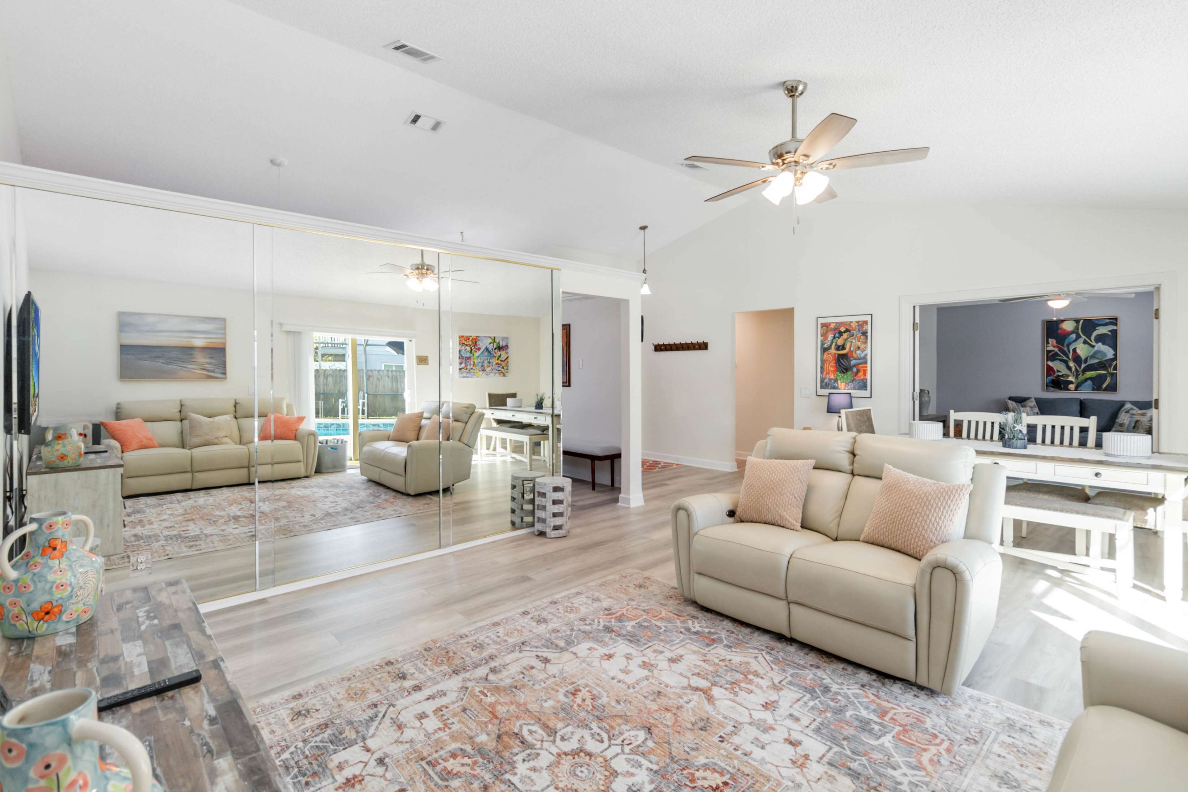 The living room features a light-colored sofa set, a large mirror wall, and a patterned area rug, with a dining area visible in the background.