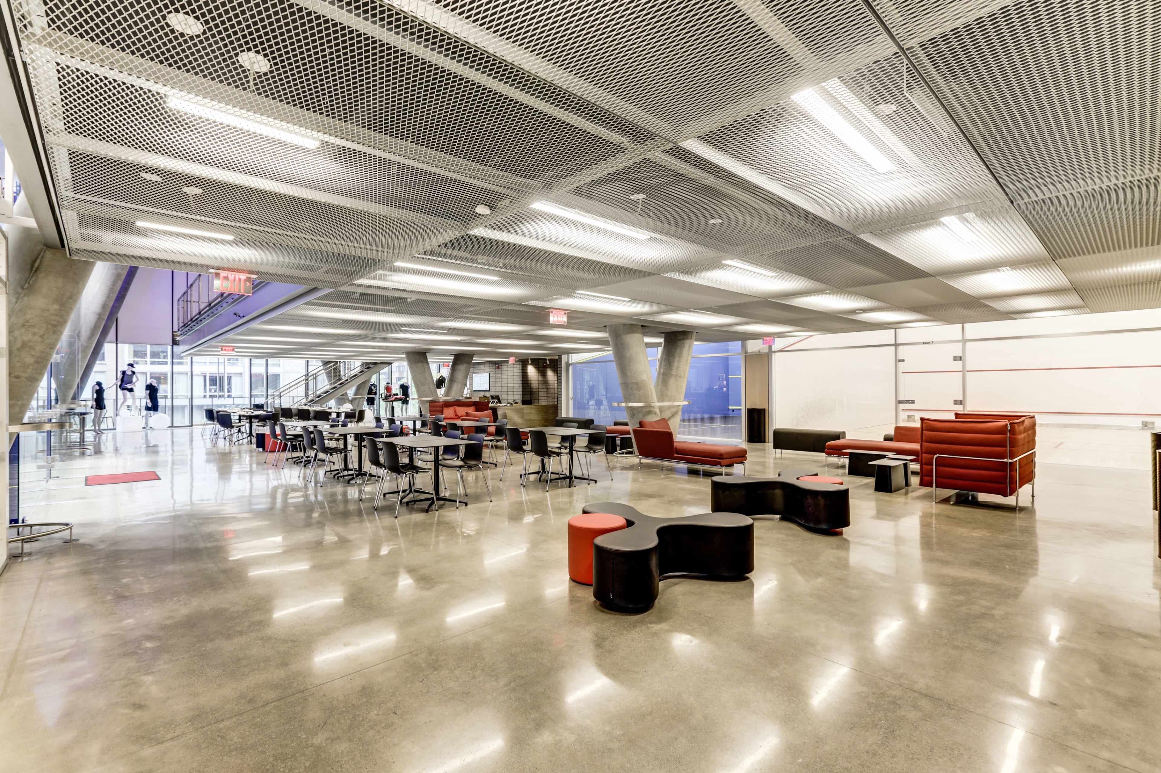 The image shows a modern, spacious indoor area with seating arrangements, tables, and a large open space under a metal grid ceiling.