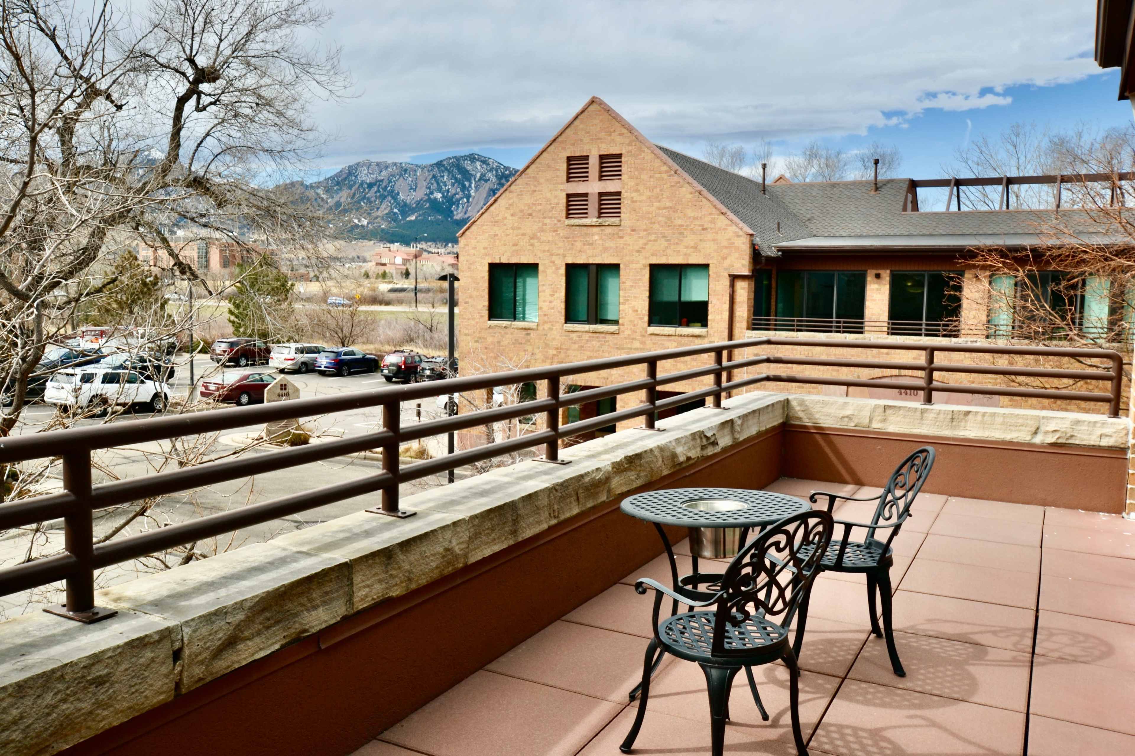 A small balcony with a stone railing features two metal chairs and a round table, overlooking a parking lot and mountains in the background.