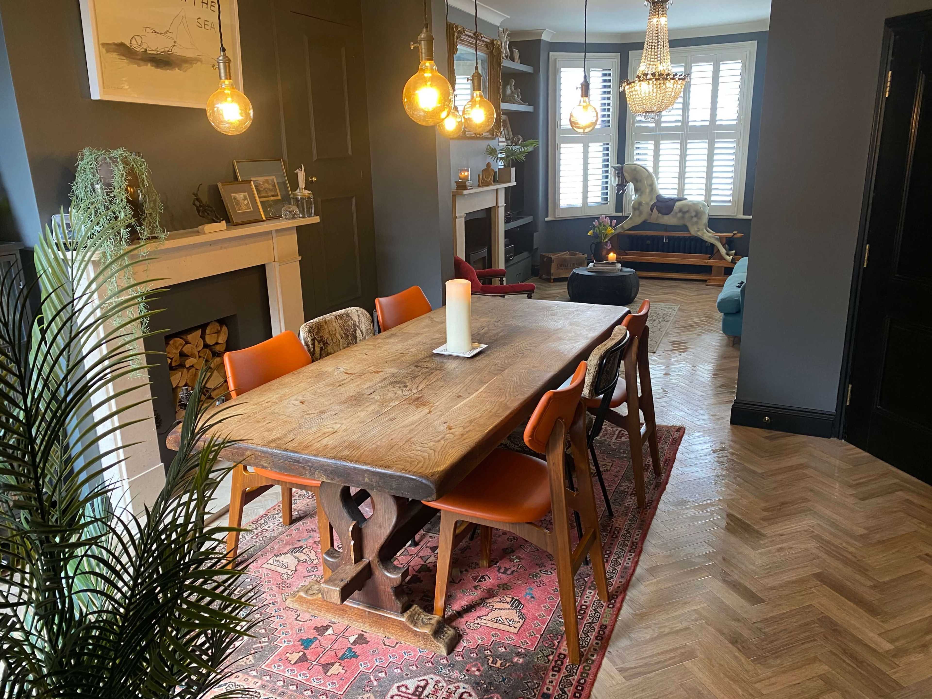 A dining room features a large wooden table surrounded by orange chairs, with a decorative rug, a fireplace, and a chandelier overhead.