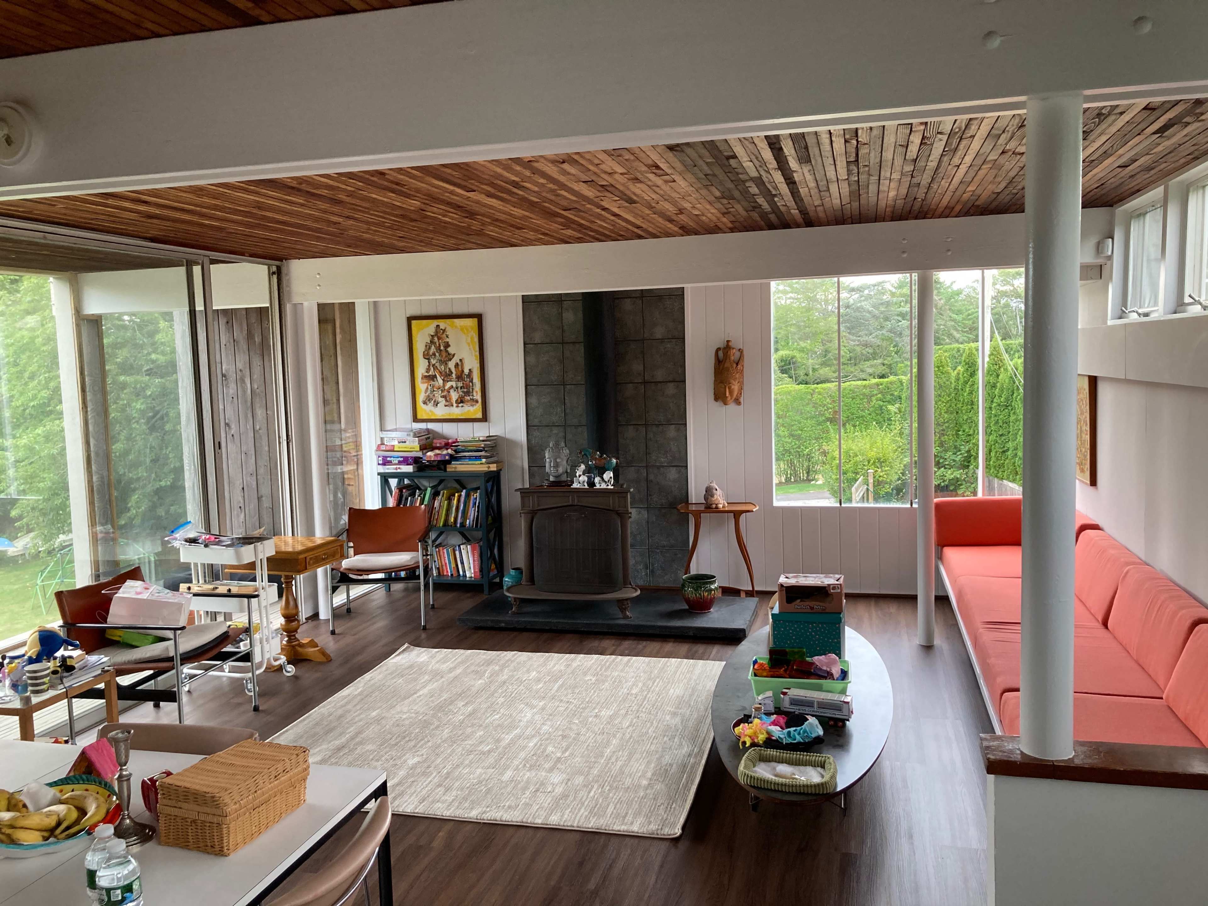 A spacious living room with wooden ceilings, a central fireplace, and large windows overlooking a green outdoor area, furnished with a red sofa and various items on tables.